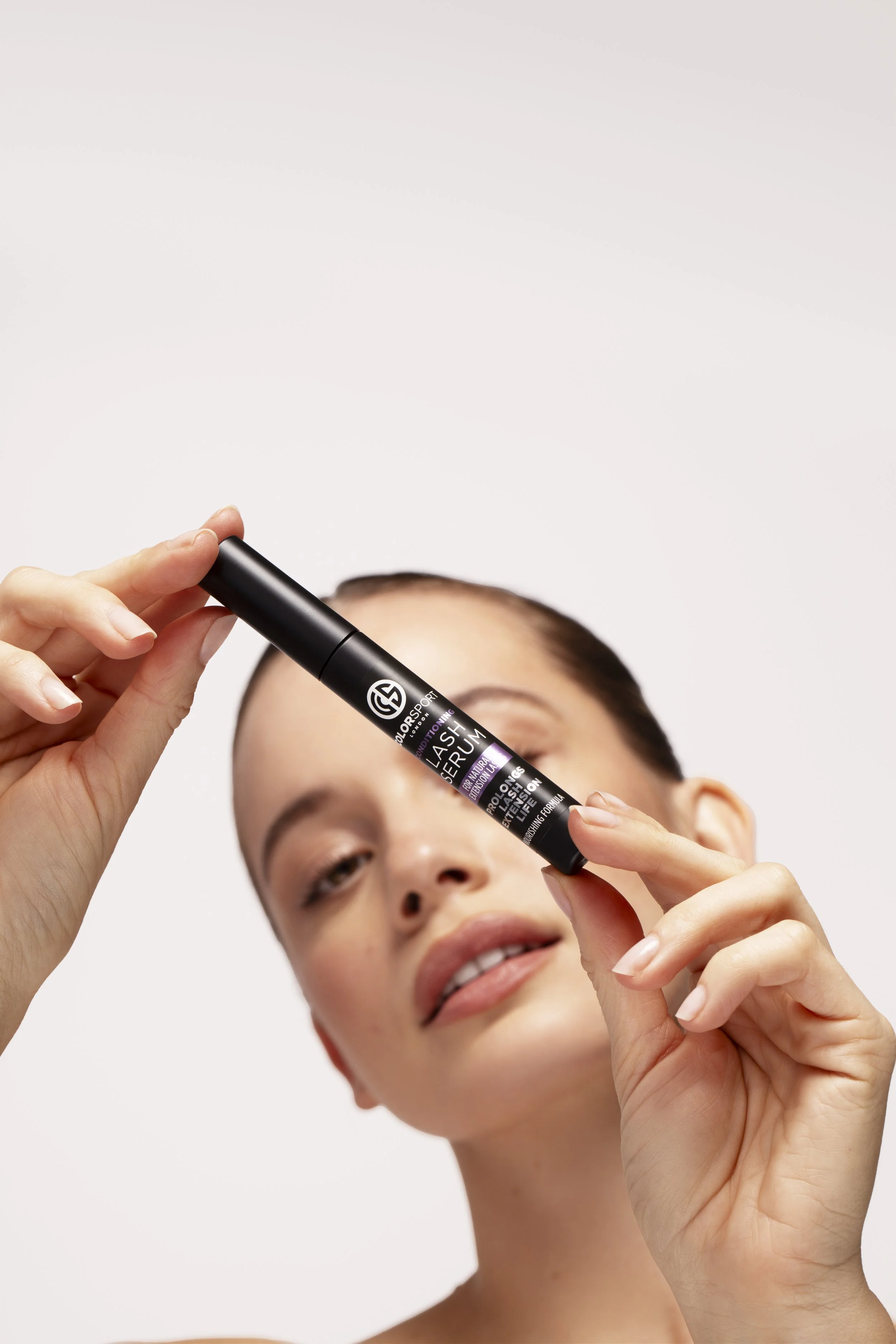 A woman holding a black tube of lash serum, looking at the camera, with a neutral expression, against a plain white background.