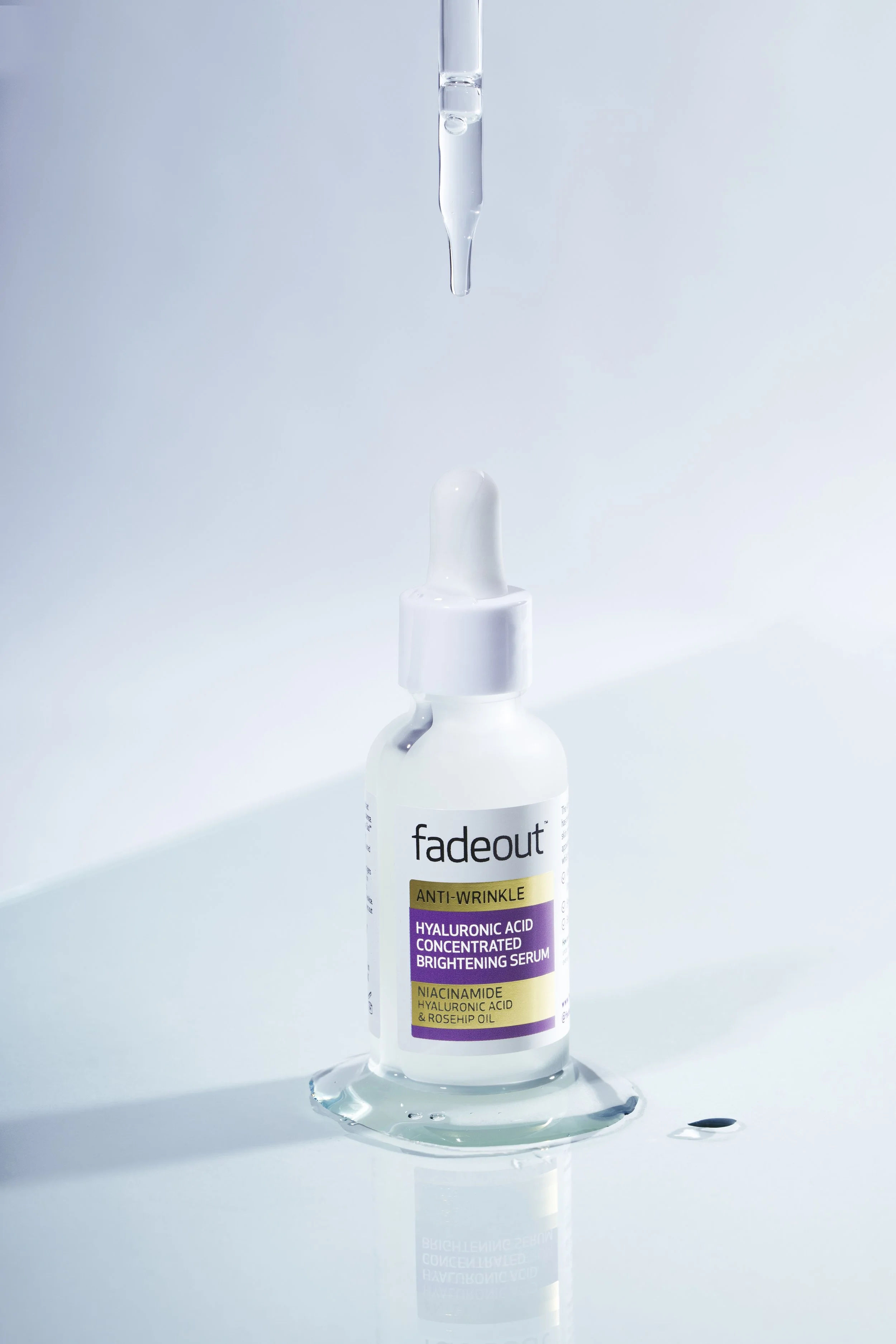 A white dropper bottle labeled 'fadeout' with 'Anti-Wrinkle Hyaluronic Acid Concentrated Brightening Serum' written on it, placed on a reflective surface with a drop of serum around the base.