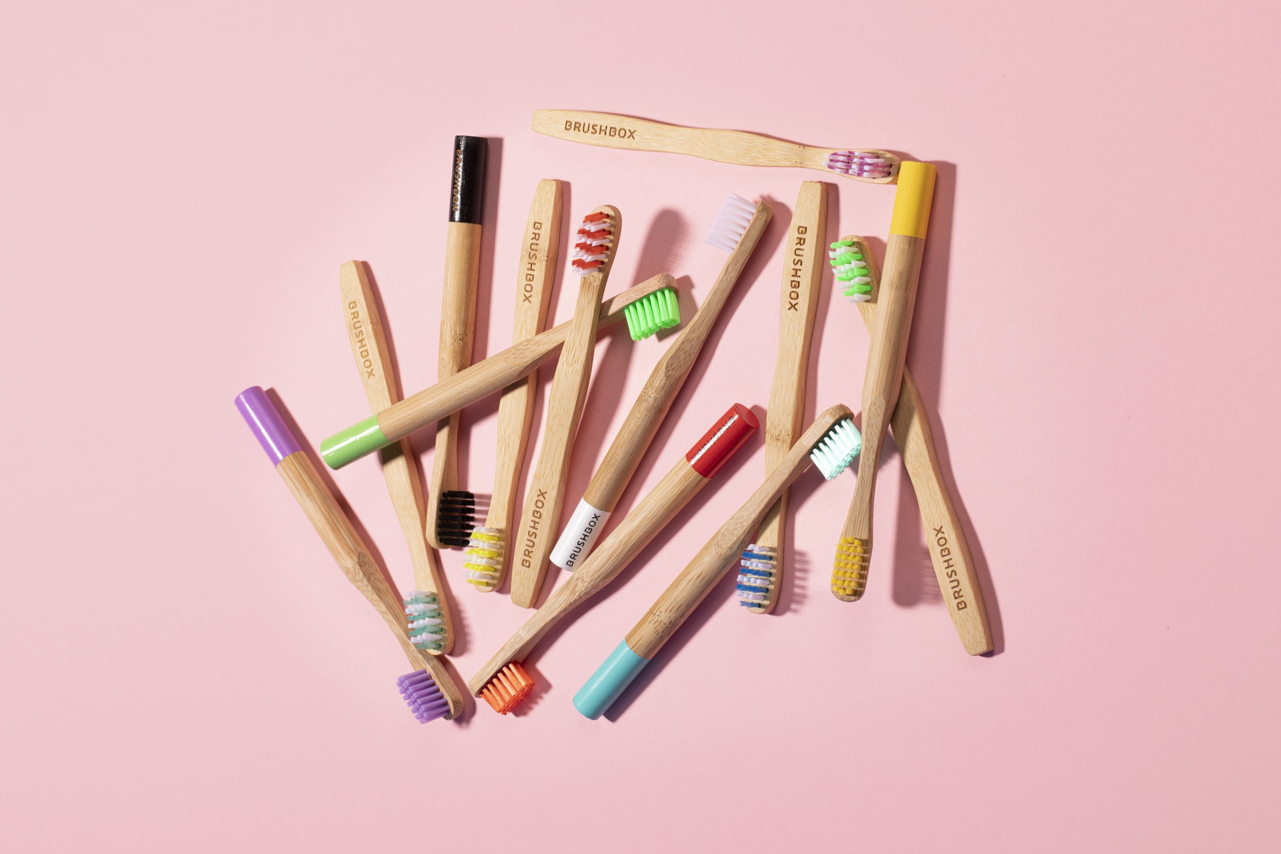 Assorted bamboo toothbrushes with colorful bristles scattered on a pink background.