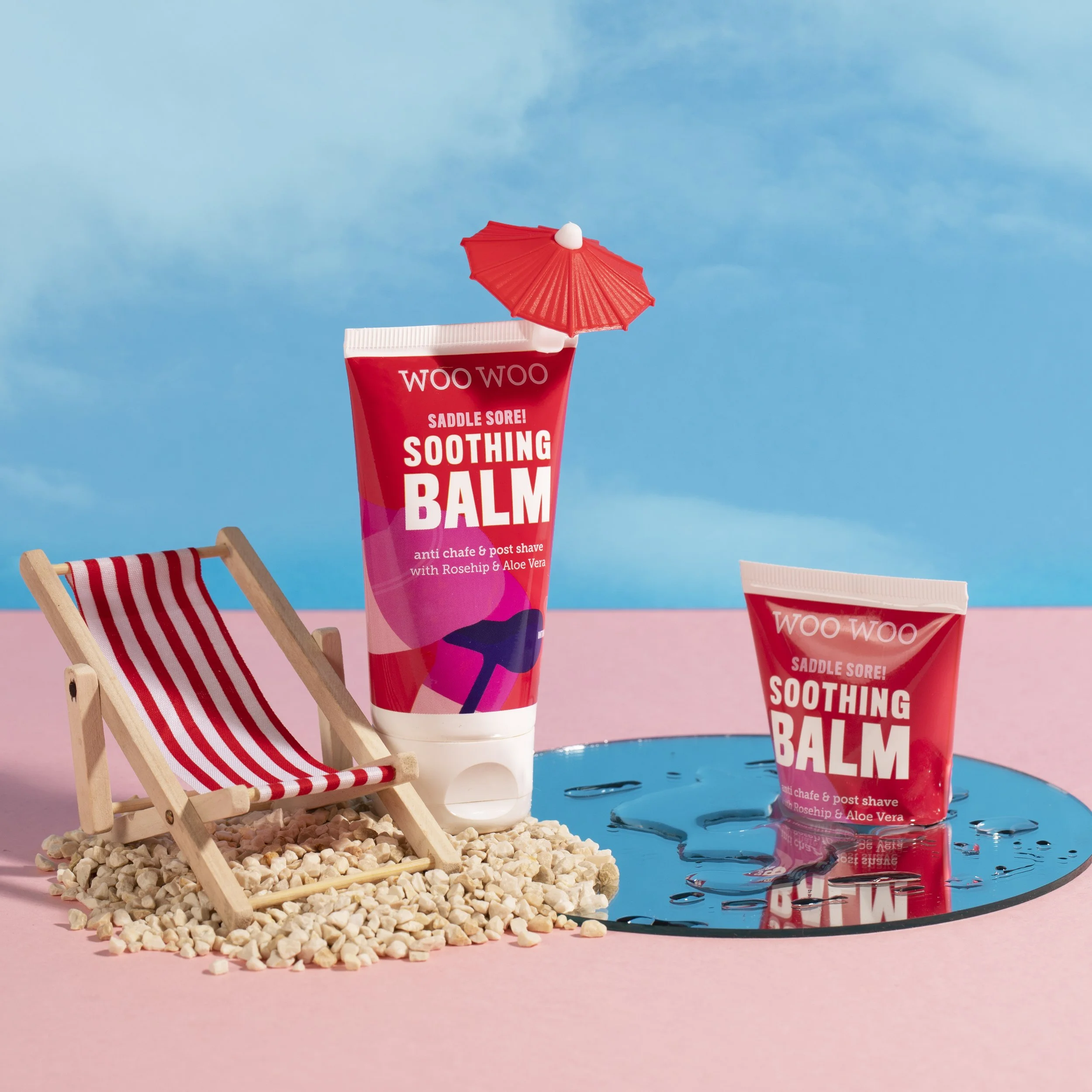 Two tubes of Woo Woo Soothing Balm with a small red cocktail umbrella. One tube is standing on a pink surface next to a miniature wooden beach chair on a bed of white pebbles. The other tube is placed on a reflective blue surface, with some blue liqu