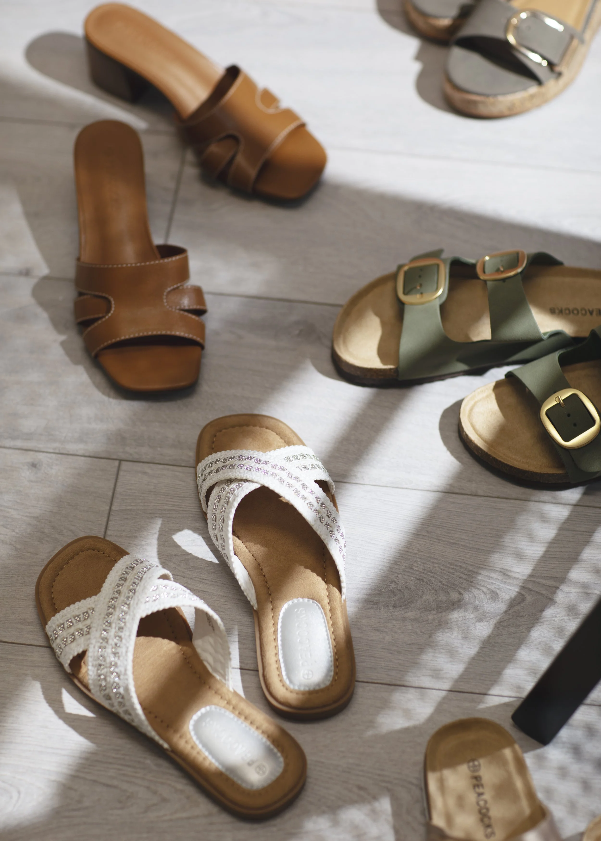 Footwear including white slip-on sandals with straps, brown slip-on sandals with straps, and green and gray sandals with buckles on a light wooden floor.