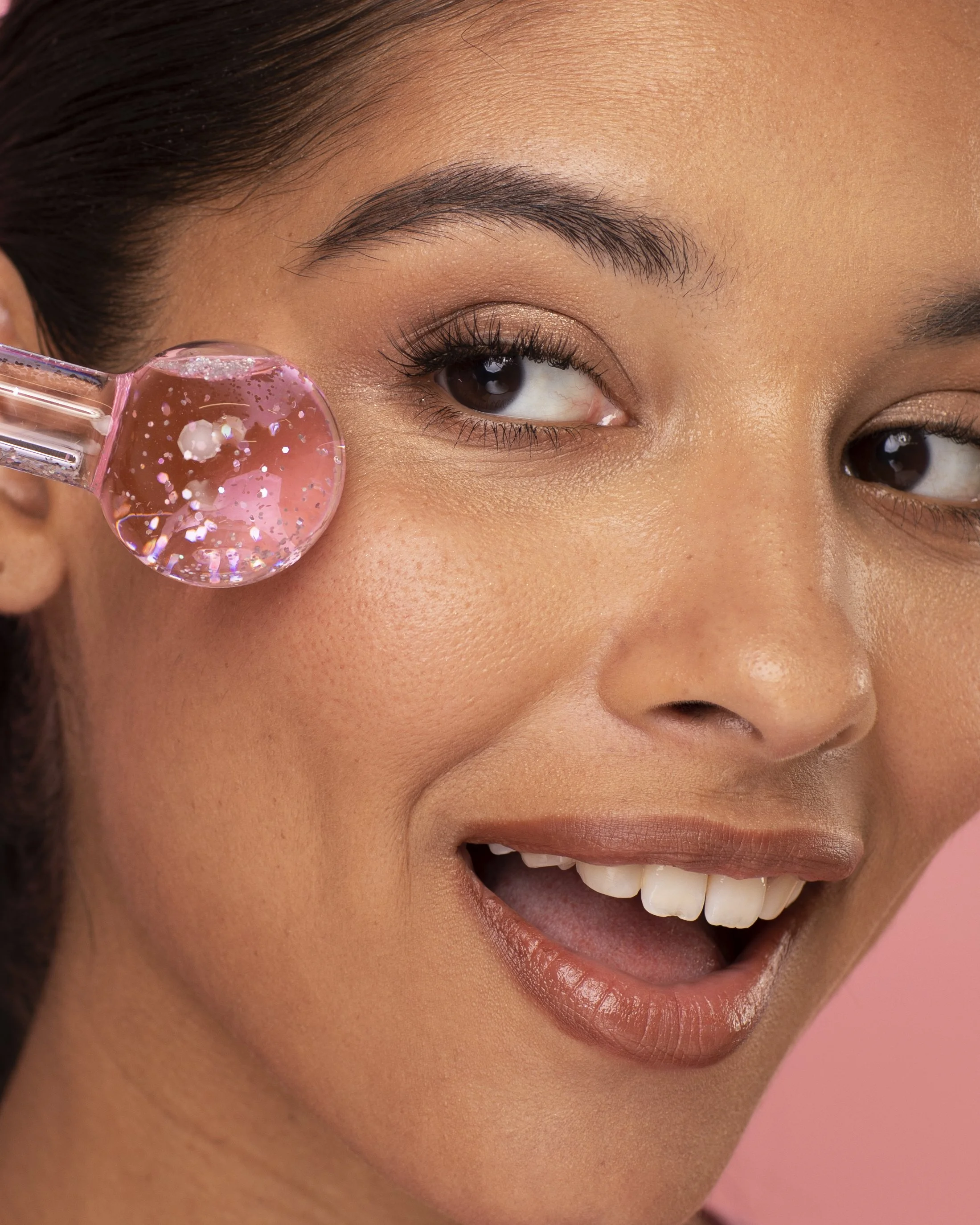 A woman with clear skin is smiling as she applies a pink facial serum with glitter particles using a dropper to her cheek.
