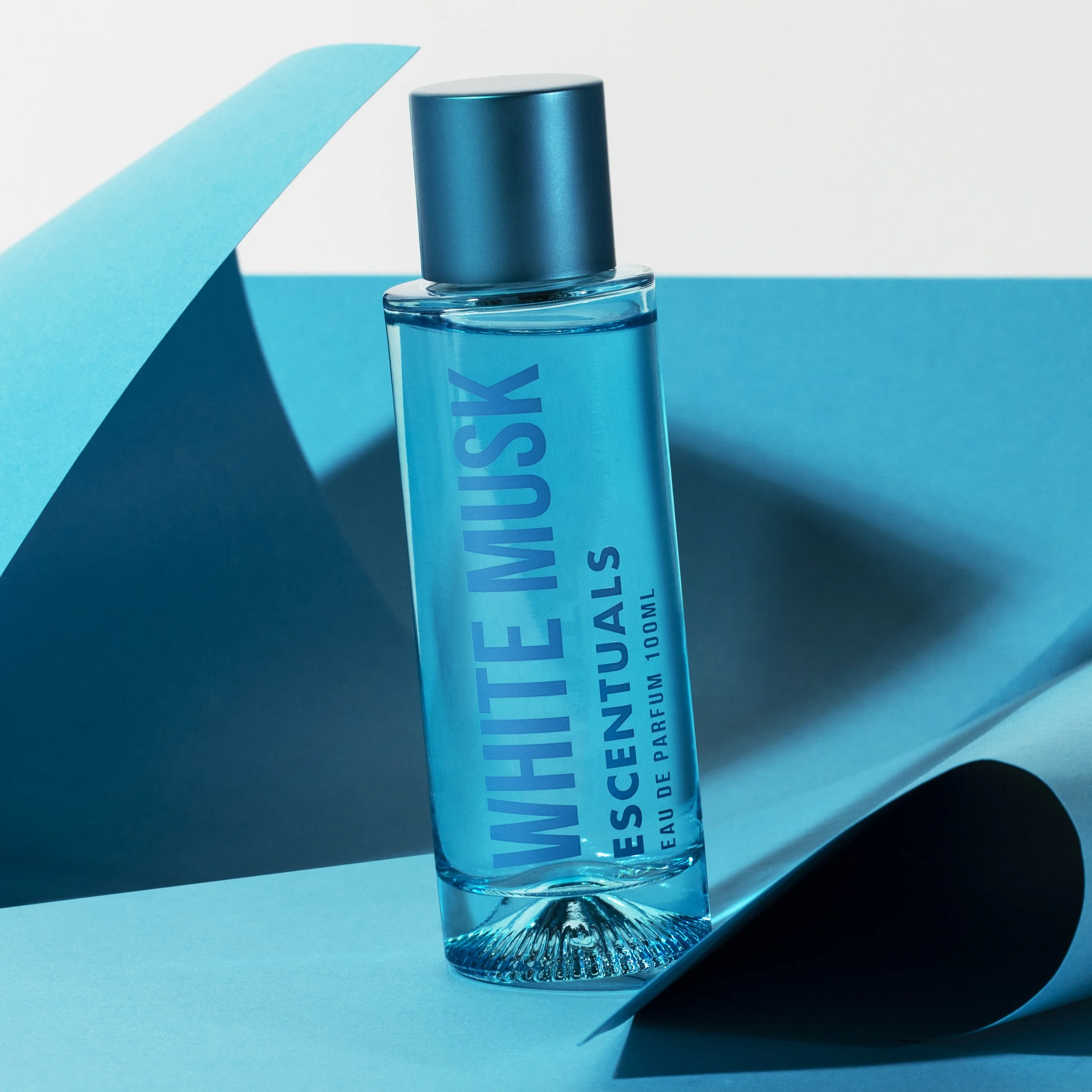A clear glass bottle of White Musk Eau de Parfum standing upright on blue and white paper sheets, with shadows cast on the background.