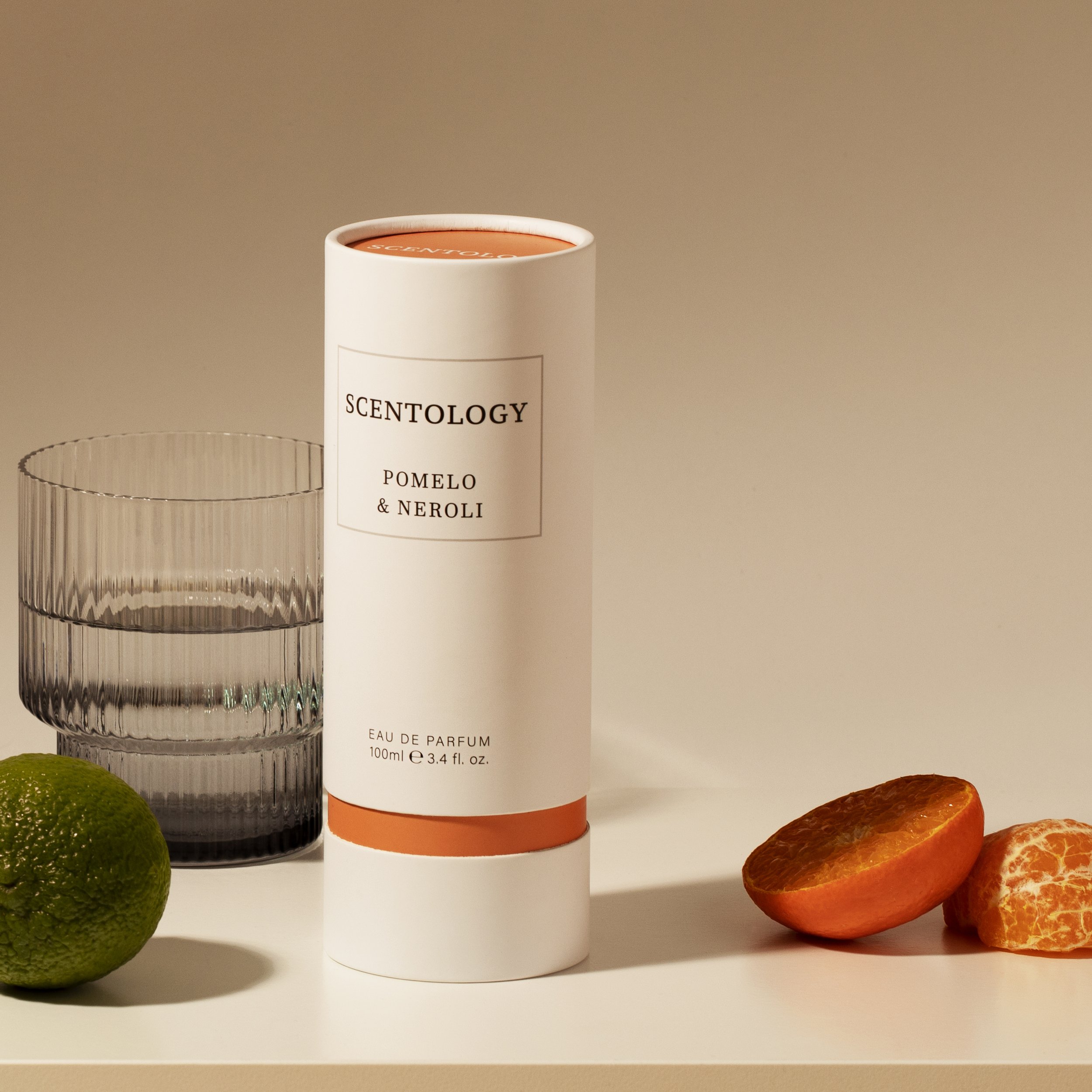 A white cylindrical perfume box labeled 'Scentology Pomelo & Neroli', a glass of water, a whole lime, and a sliced orange on a white surface against a beige background.