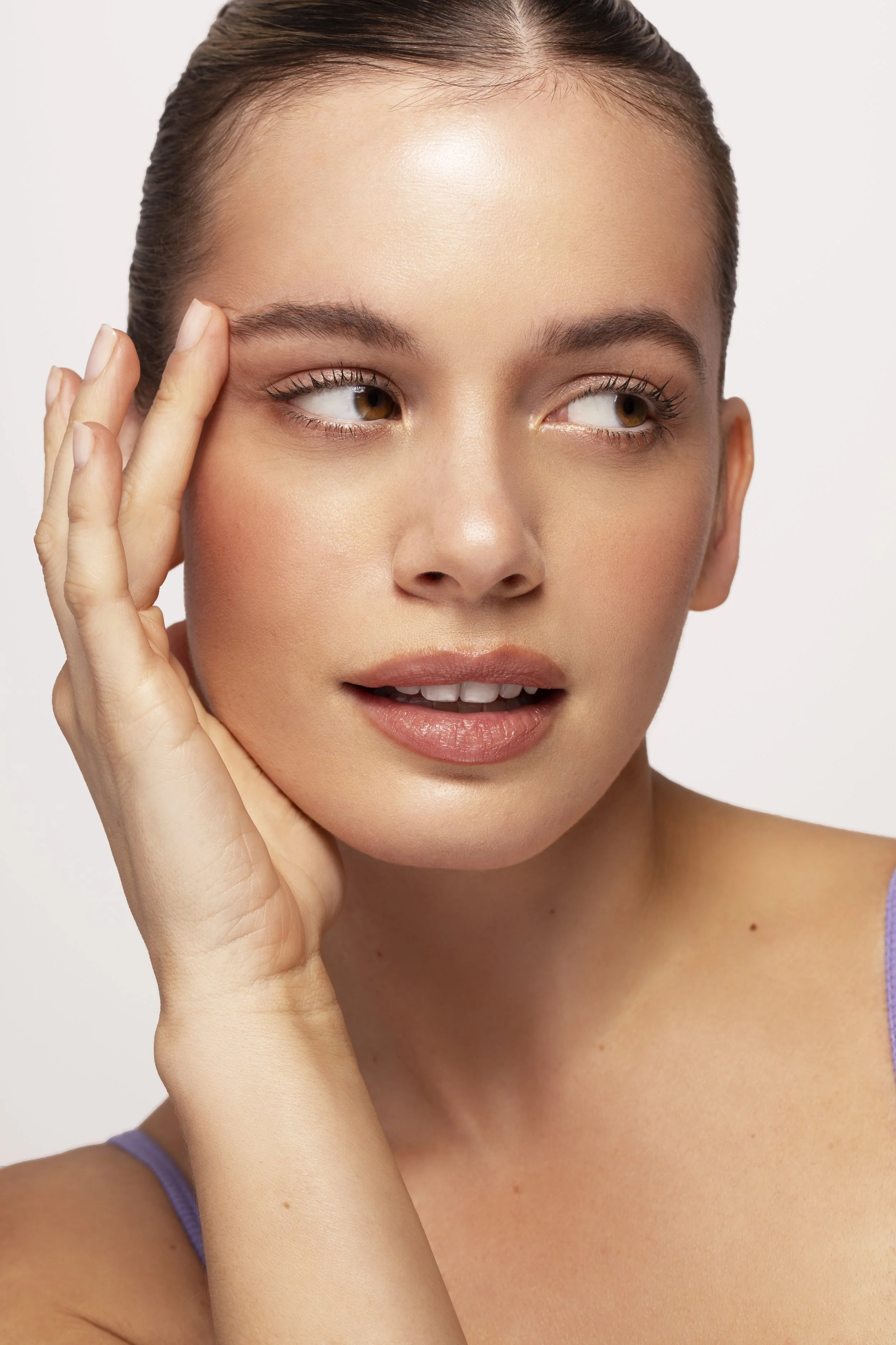 Close-up of a young woman with smooth skin, brown eyes, and brown hair, touching her face gently with her hand.