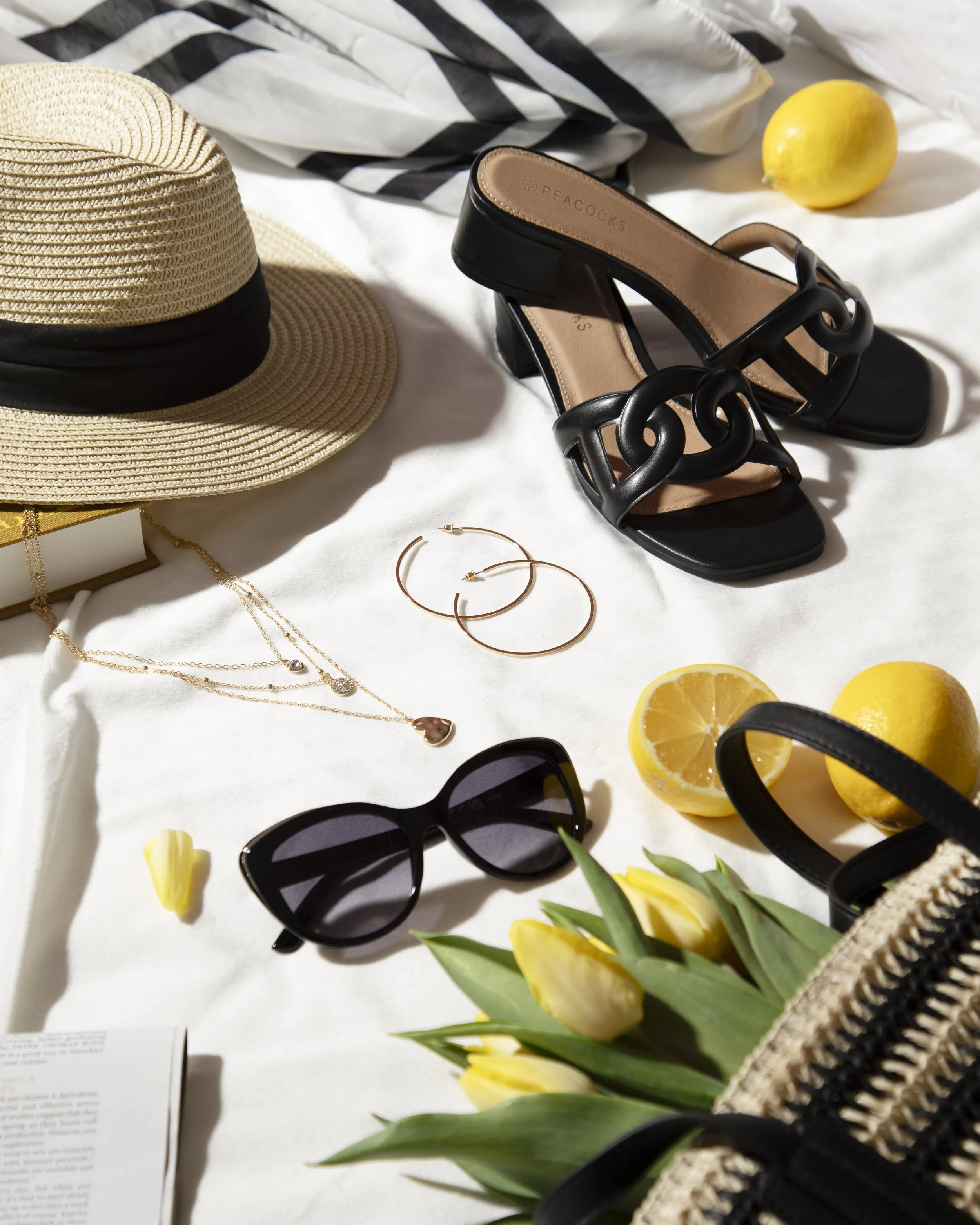 A summer-themed flat lay with a straw hat, black sandals, sunglasses, yellow lemons, tulips, jewelry, a woven bag, and an open magazine on a white surface, with a striped fabric in the background.