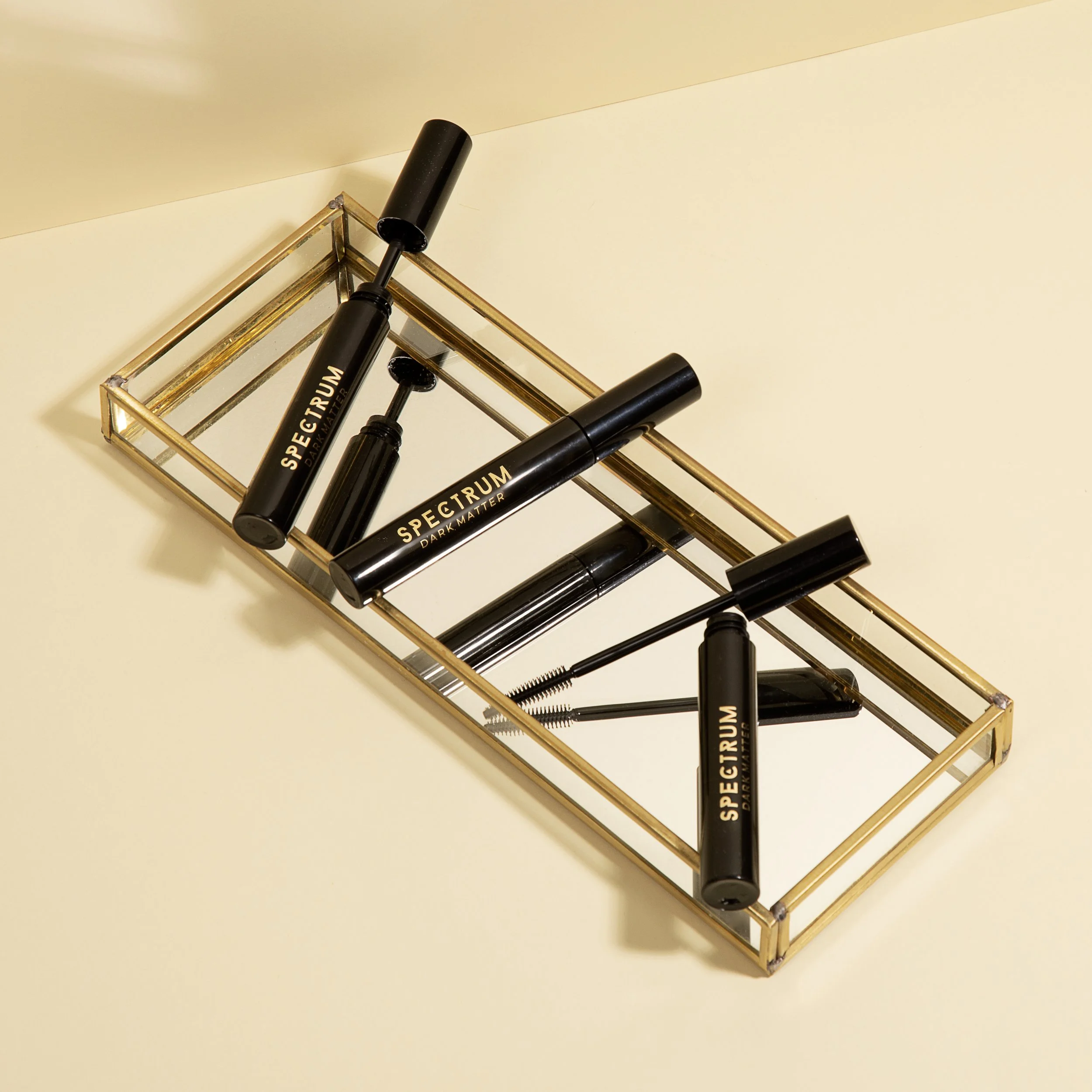 Black mascara tubes with gold lettering on a gold rectangular tray placed on a light-colored surface.
