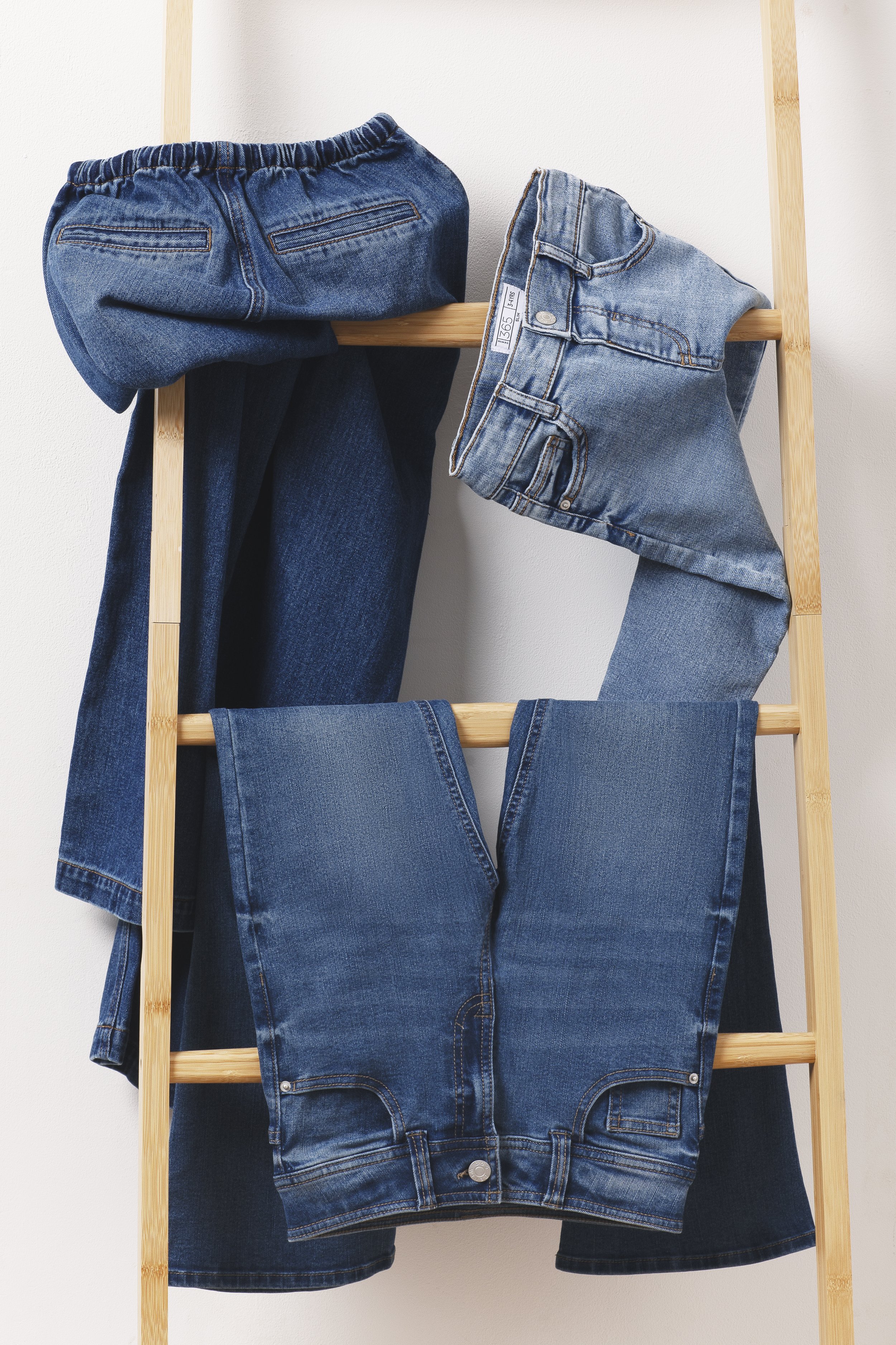 Several pairs of blue jeans draped over a wooden ladder, with a plain white background.