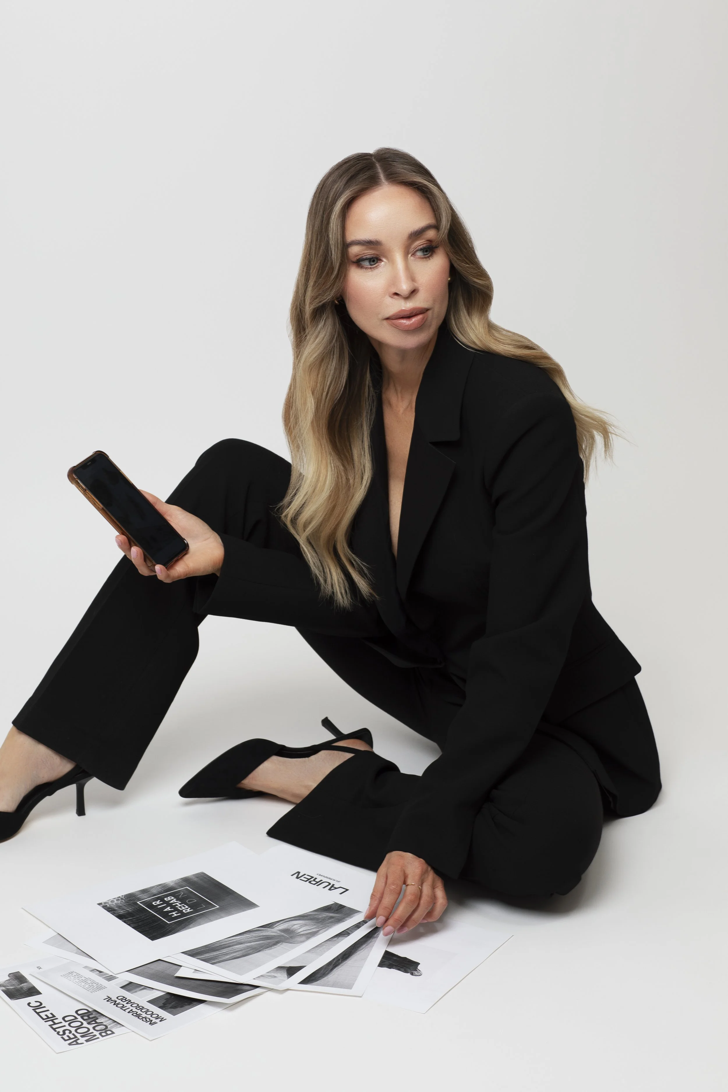 A woman in a black suit sitting cross-legged on the floor, holding a smartphone, with fashion magazines spread in front of her.