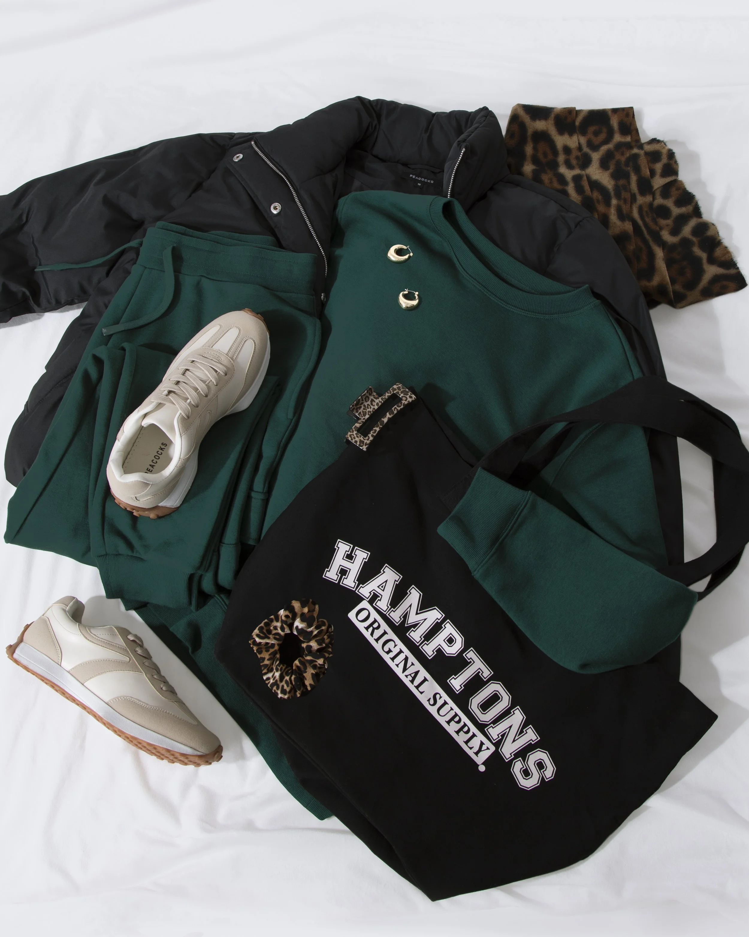 Clothing and accessories including a black puffer jacket, green sweatshirt, leopard print scarf, cream sneakers, green pants, gold earrings, and a black tote bag with white lettering.