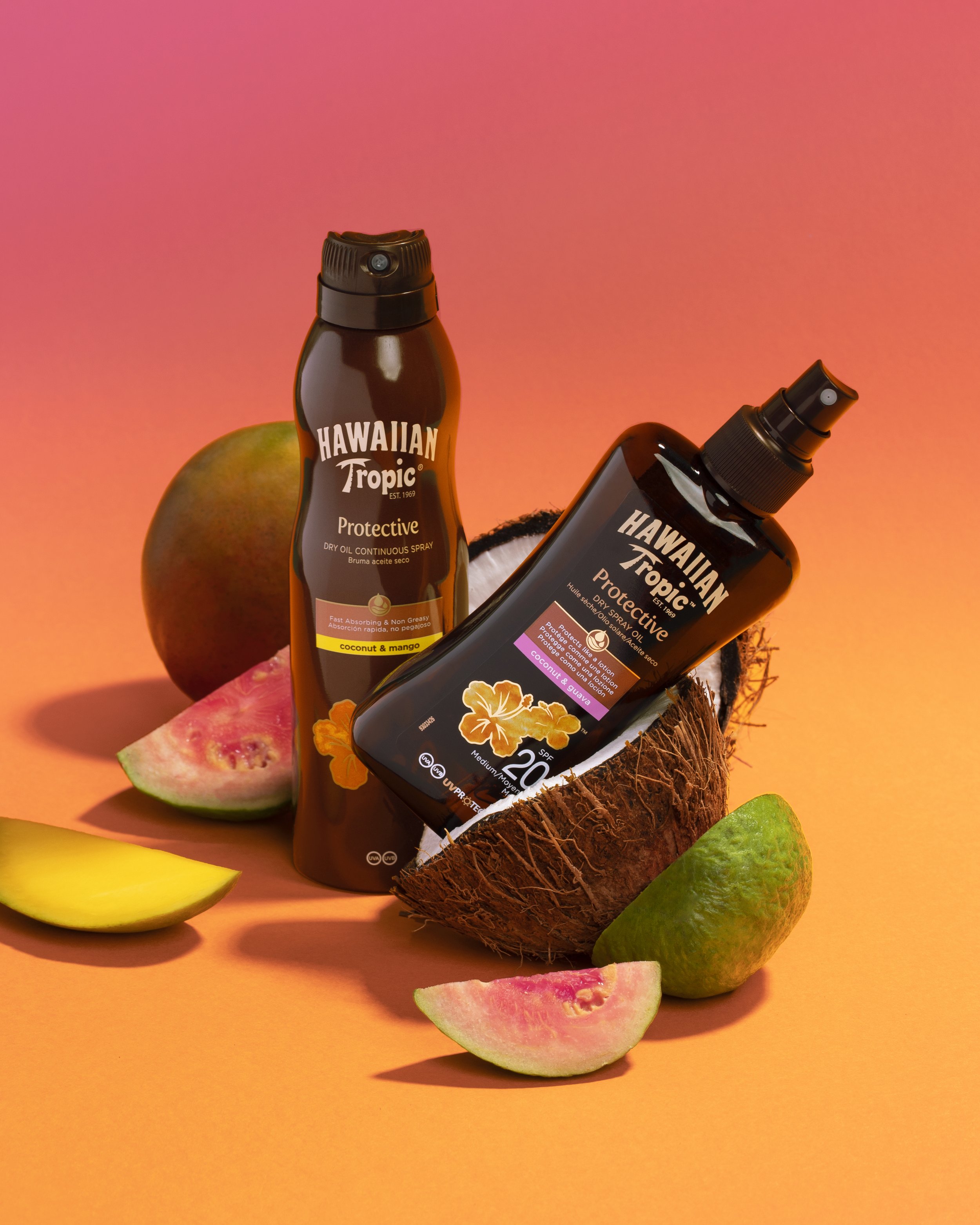 Two bottles of Hawaiian Tropic protective dry oil spray surrounded by fresh tropical fruits including a mango, guava, and slices of watermelon, set against a pink and orange background.