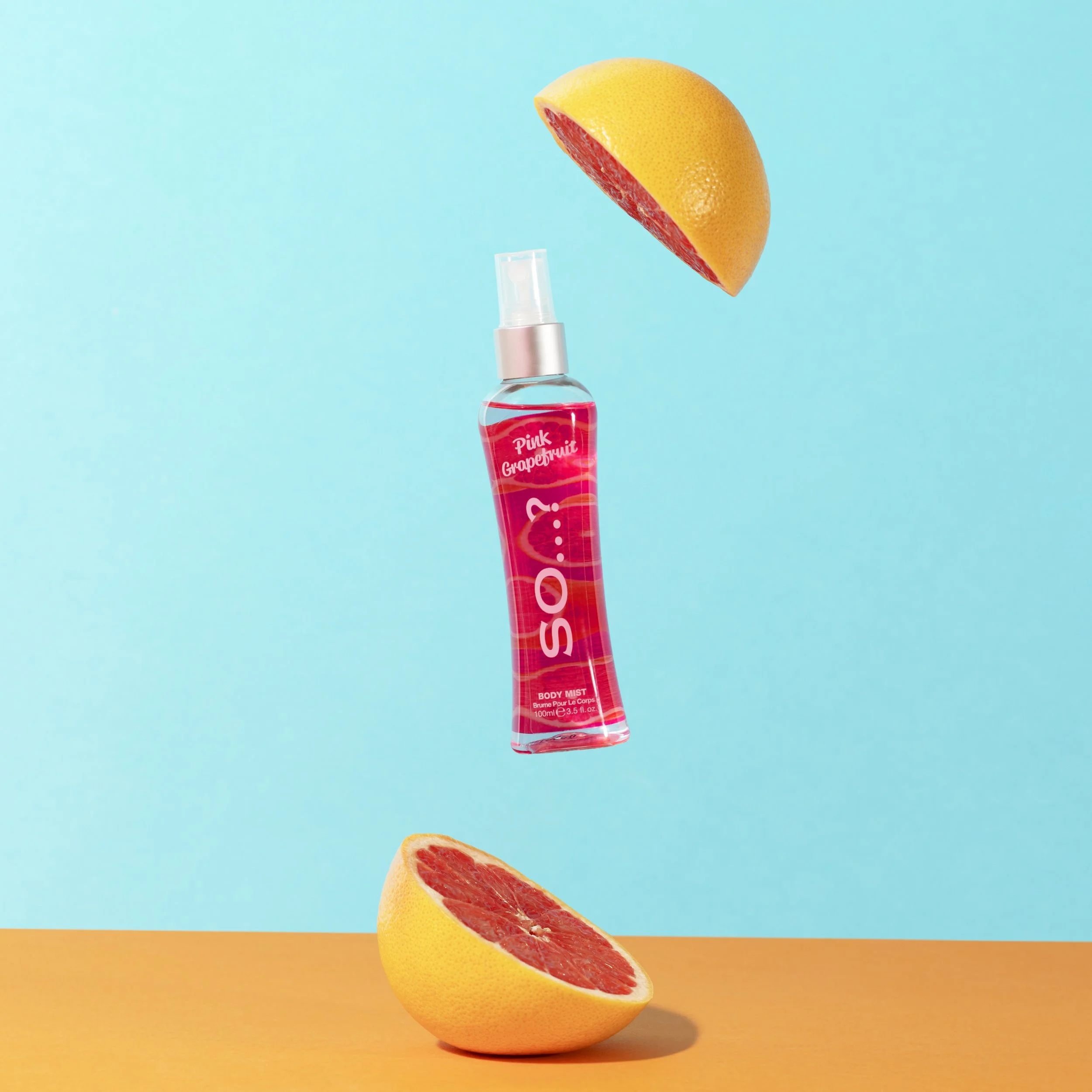 A pink grapefruit body mist spray bottle surrounded by halved pink grapefruits against a bright blue background.
