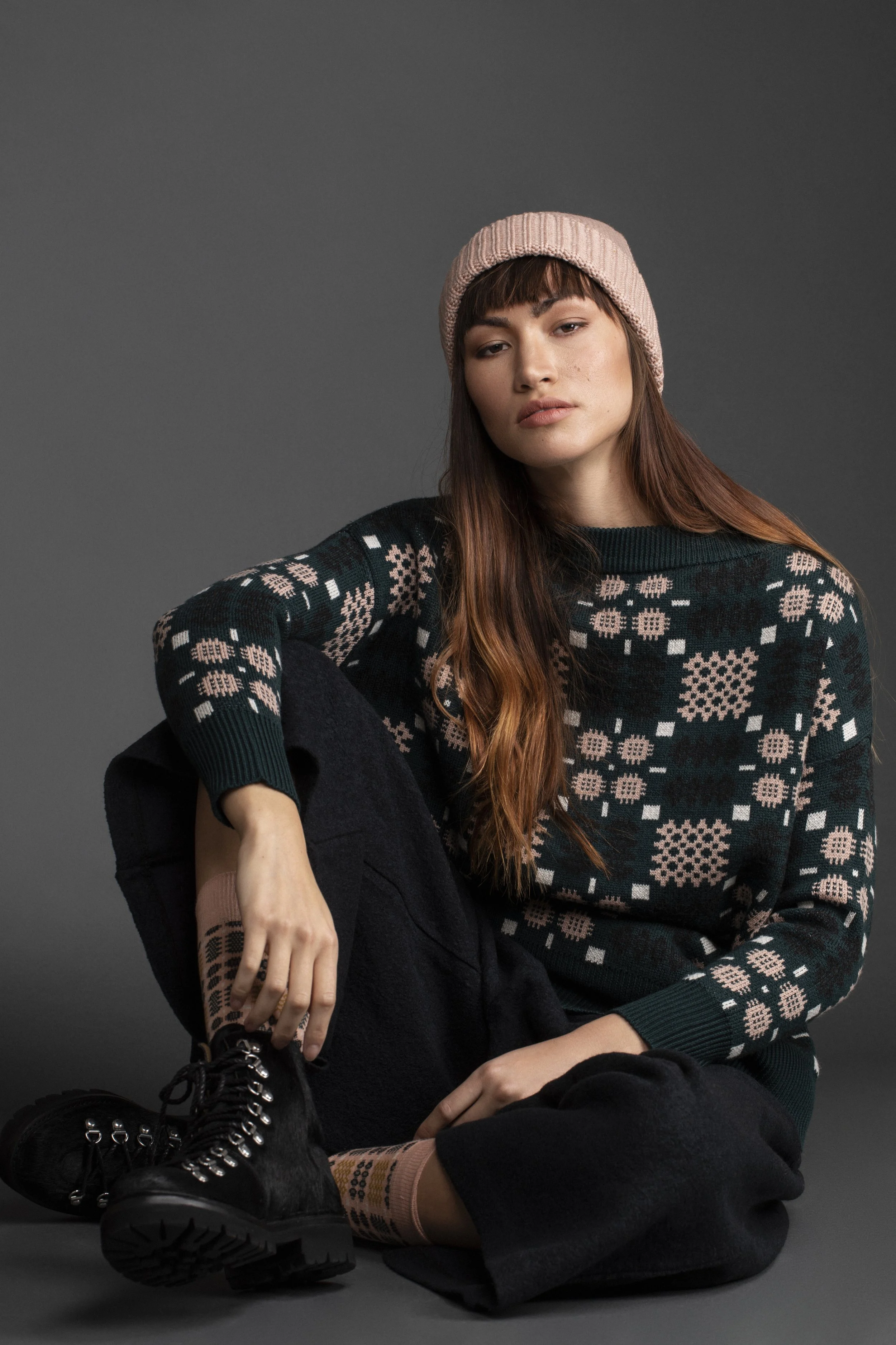 Young woman sitting on the floor against a dark gray background, wearing a pink knit beanie, a dark sweater with pink and white geometric patterns, black pants, and black lace-up boots.