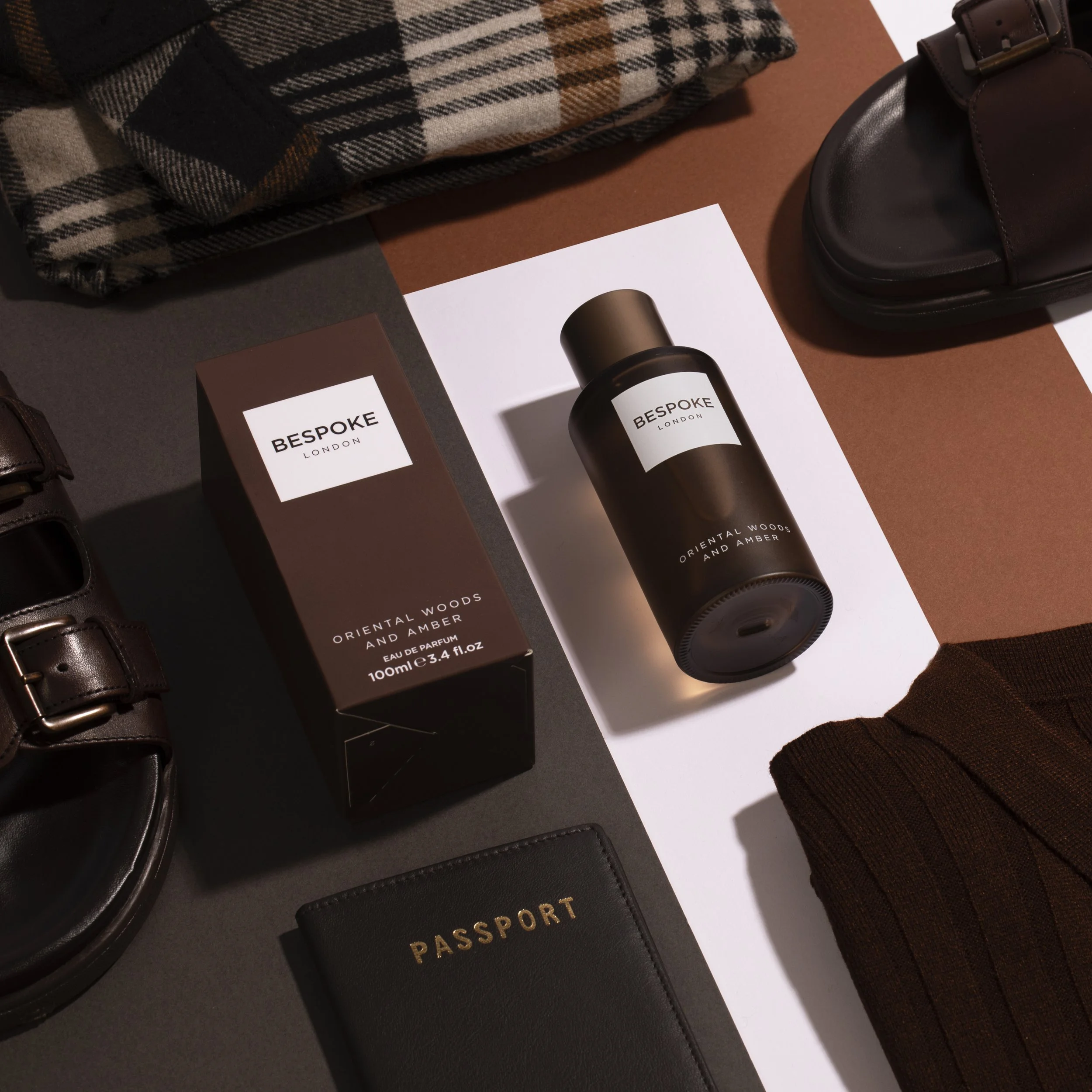 A flat lay arrangement of travel essentials including black leather shoes, a black passport holder, a brown folded scarf, a brown box labeled 'Bespoke London Oriental Woods and Amber,' a black bottle of perfume with similar branding, and a white shee