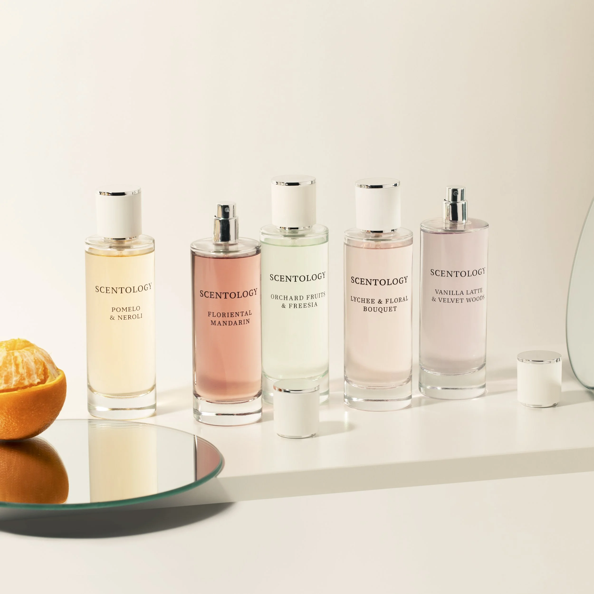 Five bottles of scented perfume on a white surface, labeled with different fragrance names, next to a halved orange and a small round mirror.