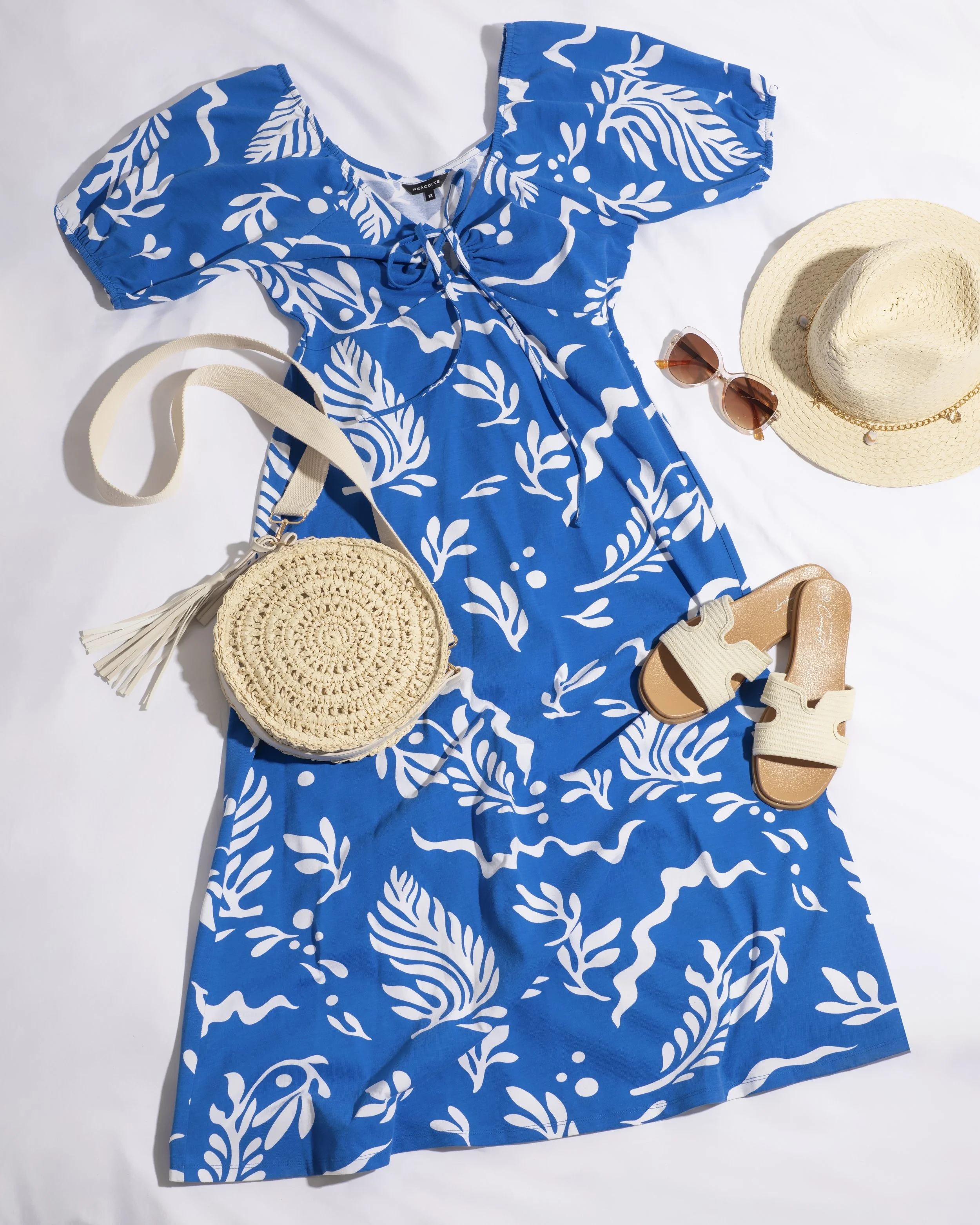 Flat lay of a summer outfit with a blue and white floral dress, beige sandals, a straw hat, sunglasses, a round woven bag with a tassel, and a straw sun hat on a white surface.