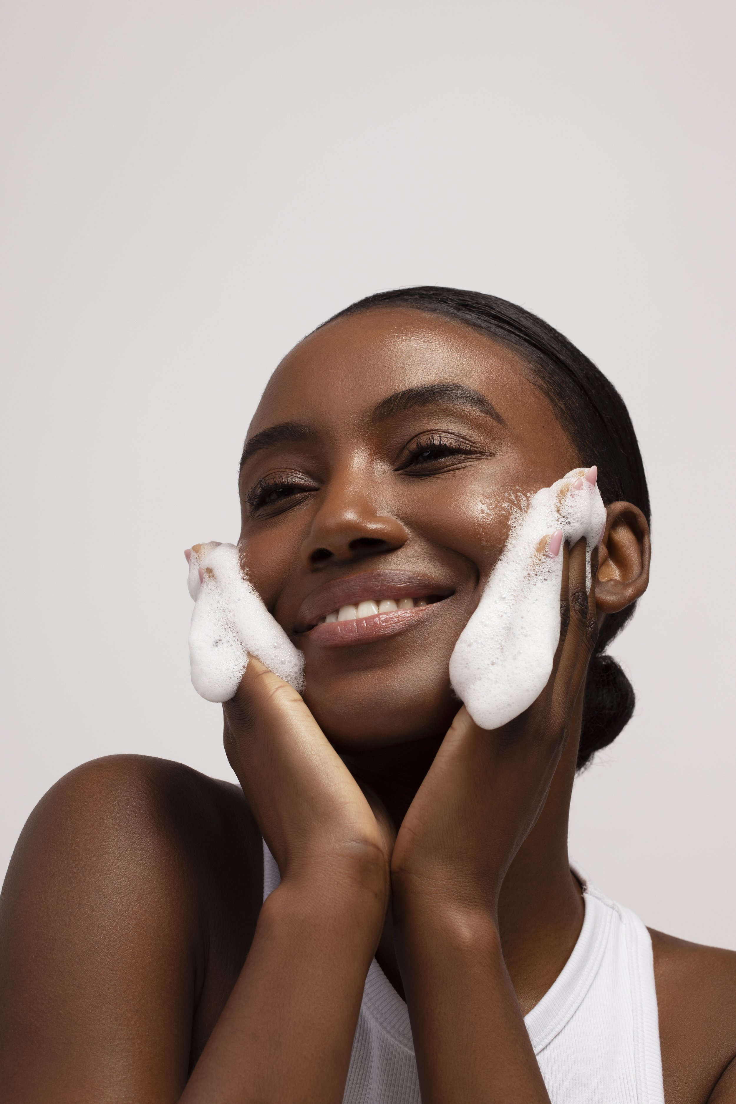 A woman smiling while washing her face with soap and foam.