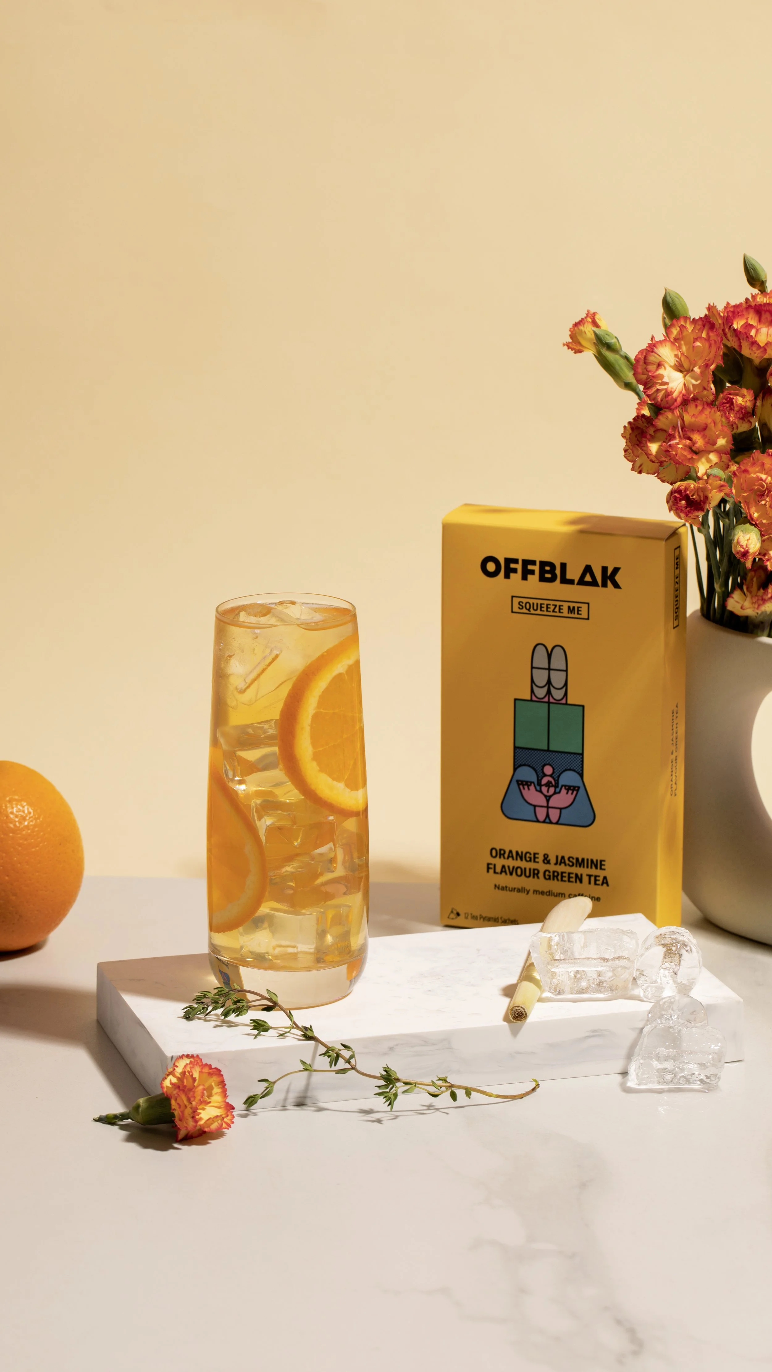 A glass of orange and jasmine flavored green tea with ice and orange slices, a box of OFFBLAK tea, a vase of pink and orange carnations, an orange fruit, a sprig of thyme, and ice cubes on a white marble surface against a beige background.