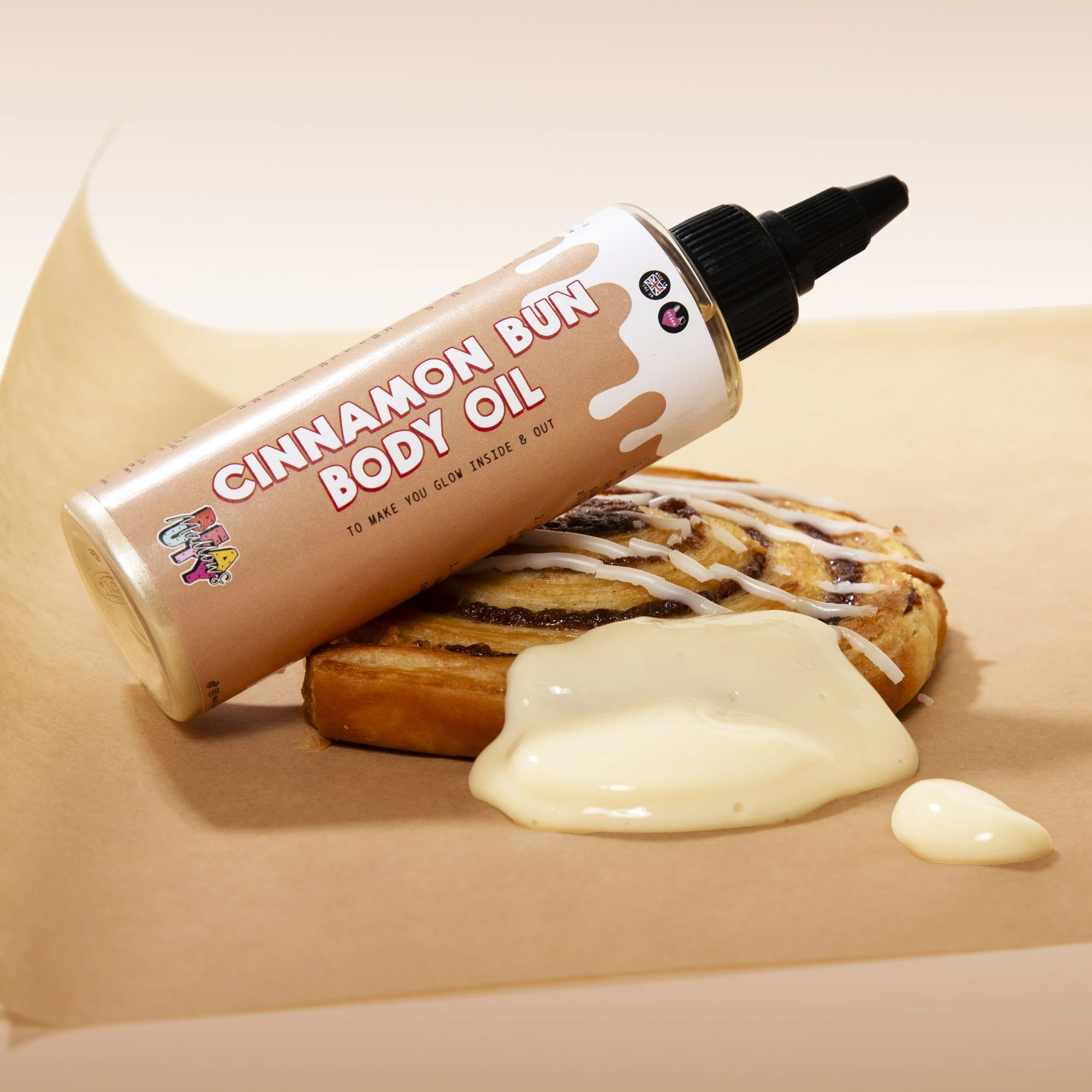 A cinnamon bun with icing, a dollop of white body oil, and a bottle labeled 'Cinnamon Bun Body Oil' resting on the bun.