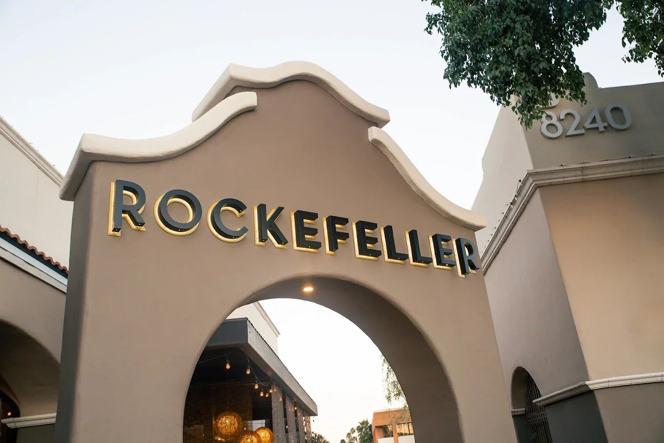 The entrance gate of Rockefeller with a sign displaying the name and the number 8240, featuring a curved arch design, situated in a shopping or commercial area.