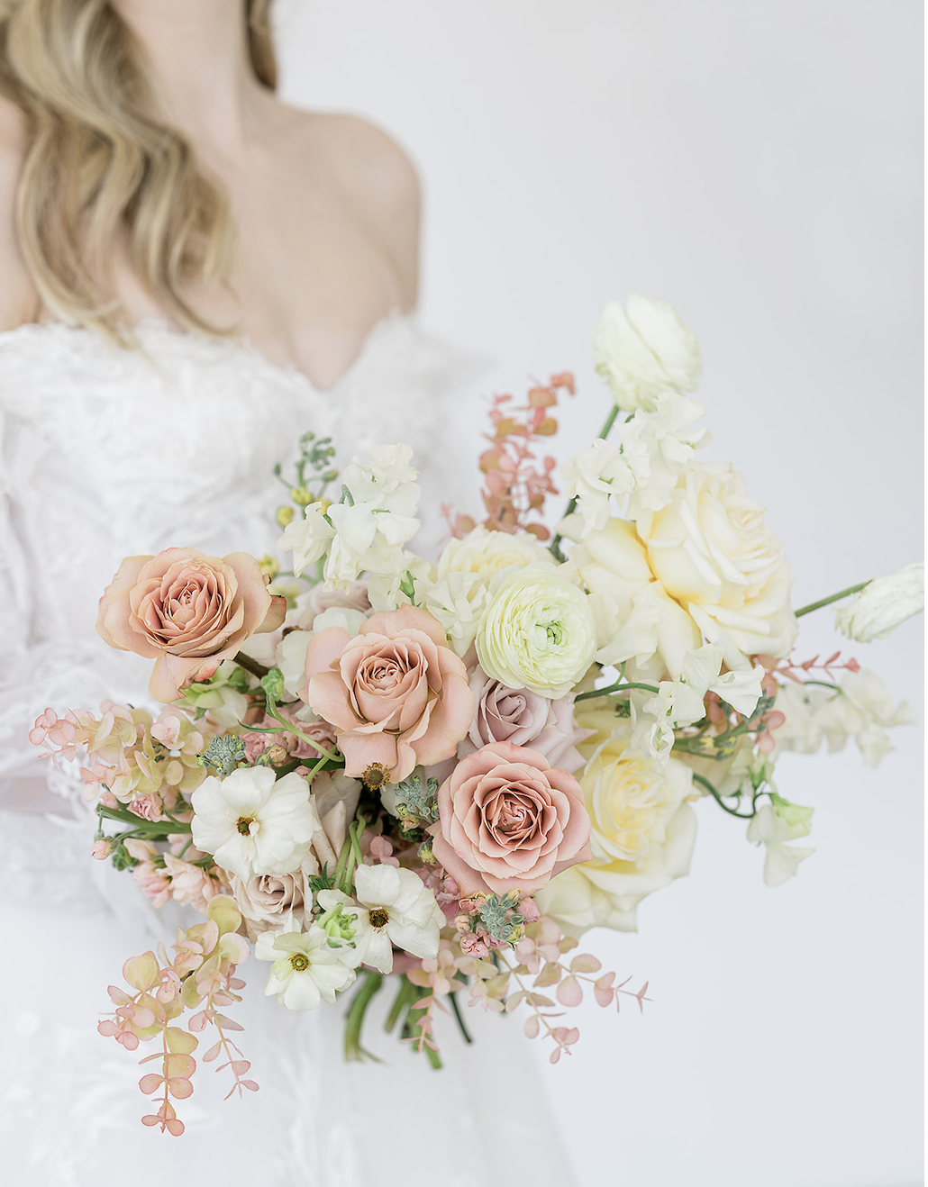 A woman in a white wedding dress holds a large bouquet of pale pink, cream, and white flowers, including roses and other mixed blooms.