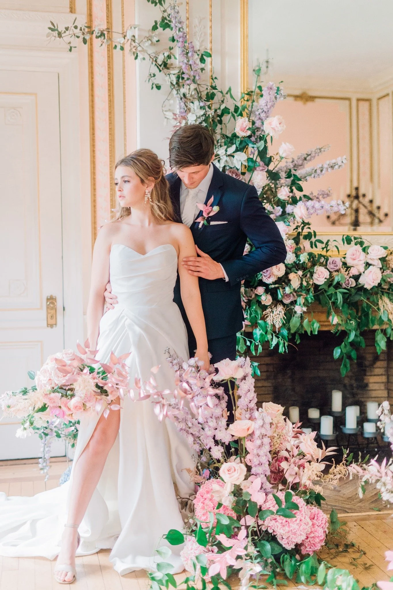 Bride and groom standing close together in a decorated wedding ceremony space with pink and purple flowers, greenery, and candles in the background.