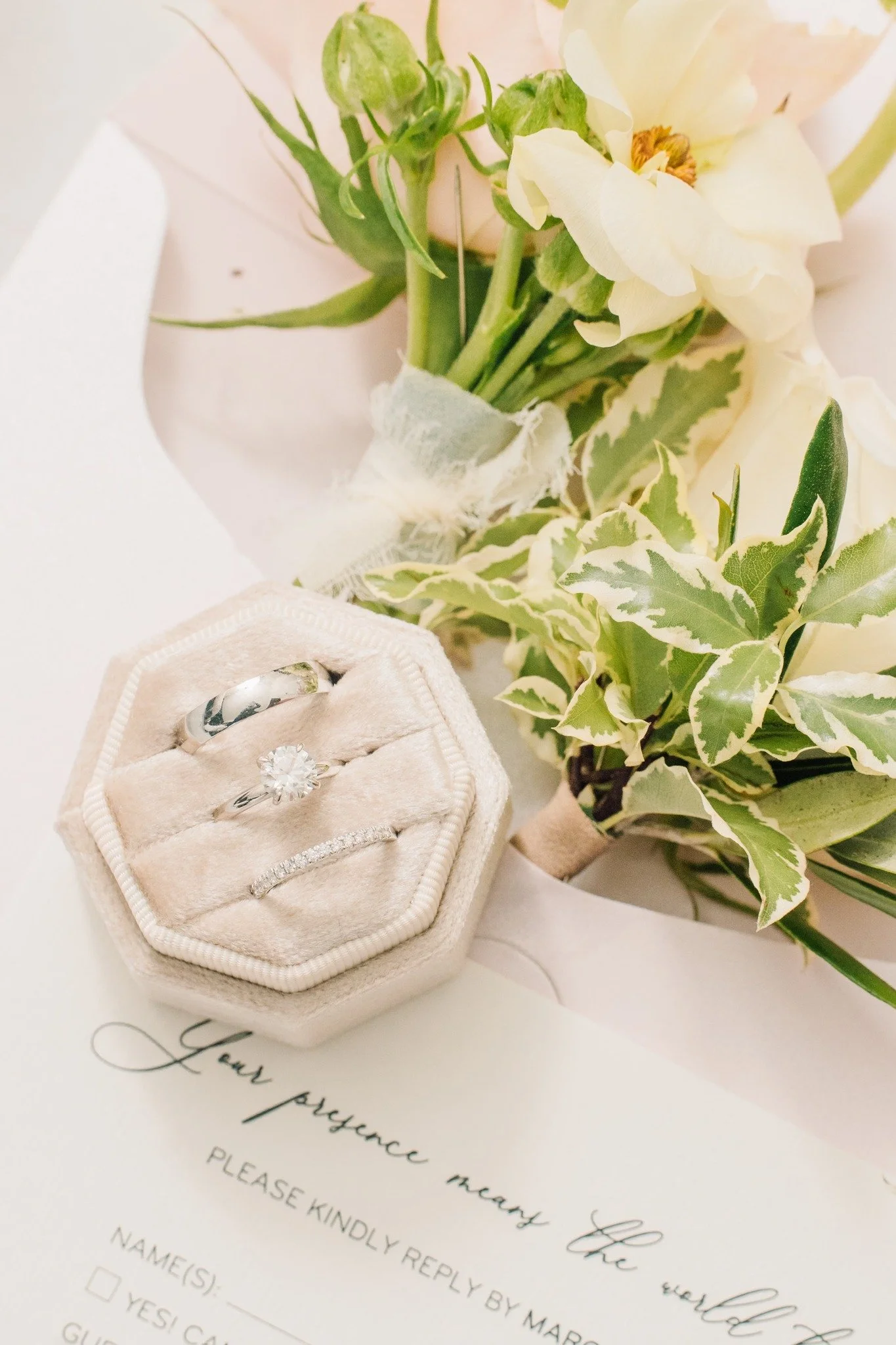 A wedding ring set in a plush, light-colored ring box with a diamond engagement ring and a wedding band, alongside a bouquet of white and green flowers on a wedding invitation.