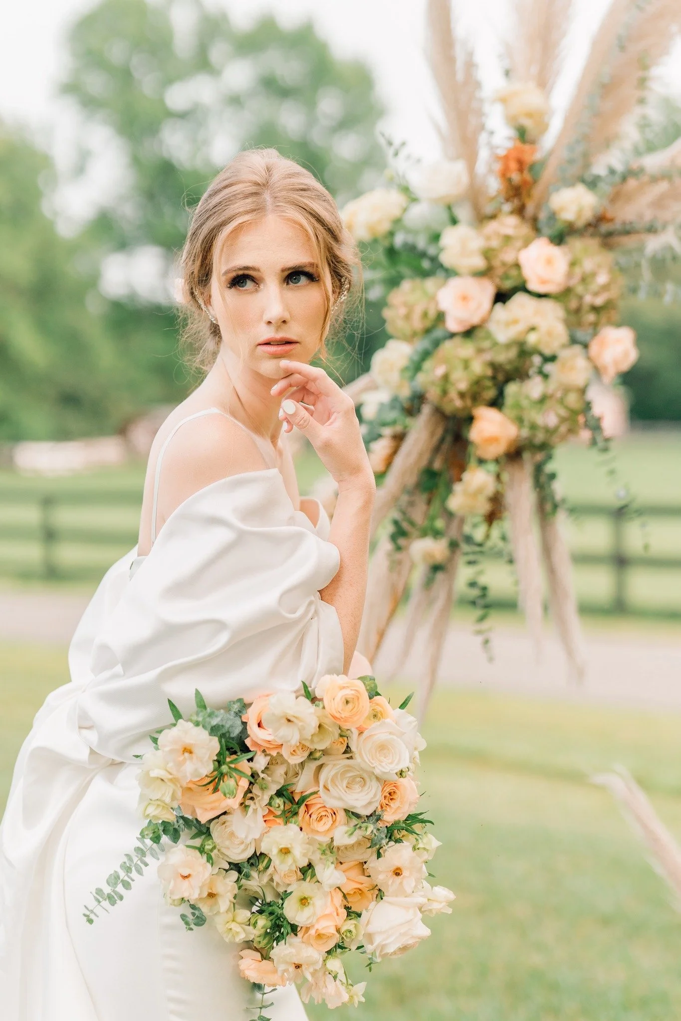 A woman in a white dress holding a cascading bouquet of peach and white roses, standing outdoors near a floral wedding arch decorated with roses and greenery.
