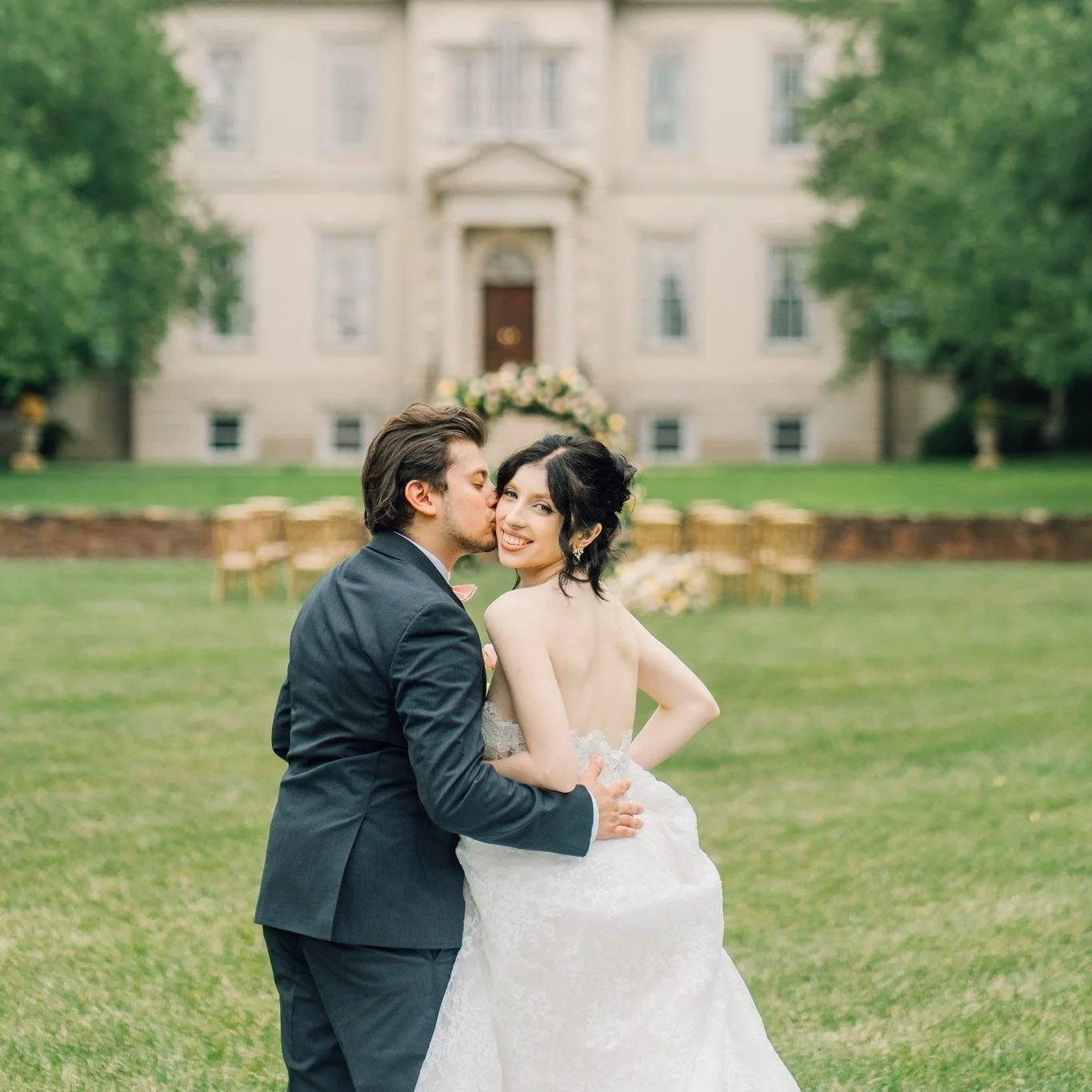 A newlywed couple in wedding attire on a green lawn in front of a large historic building. The groom is kissing the bride on the cheek, and she is smiling at the camera.