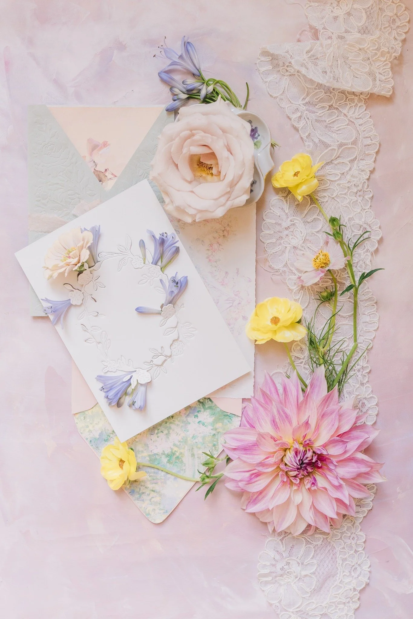 Flat lay of floral greeting card, lace trim, and fresh flowers on pink background.
