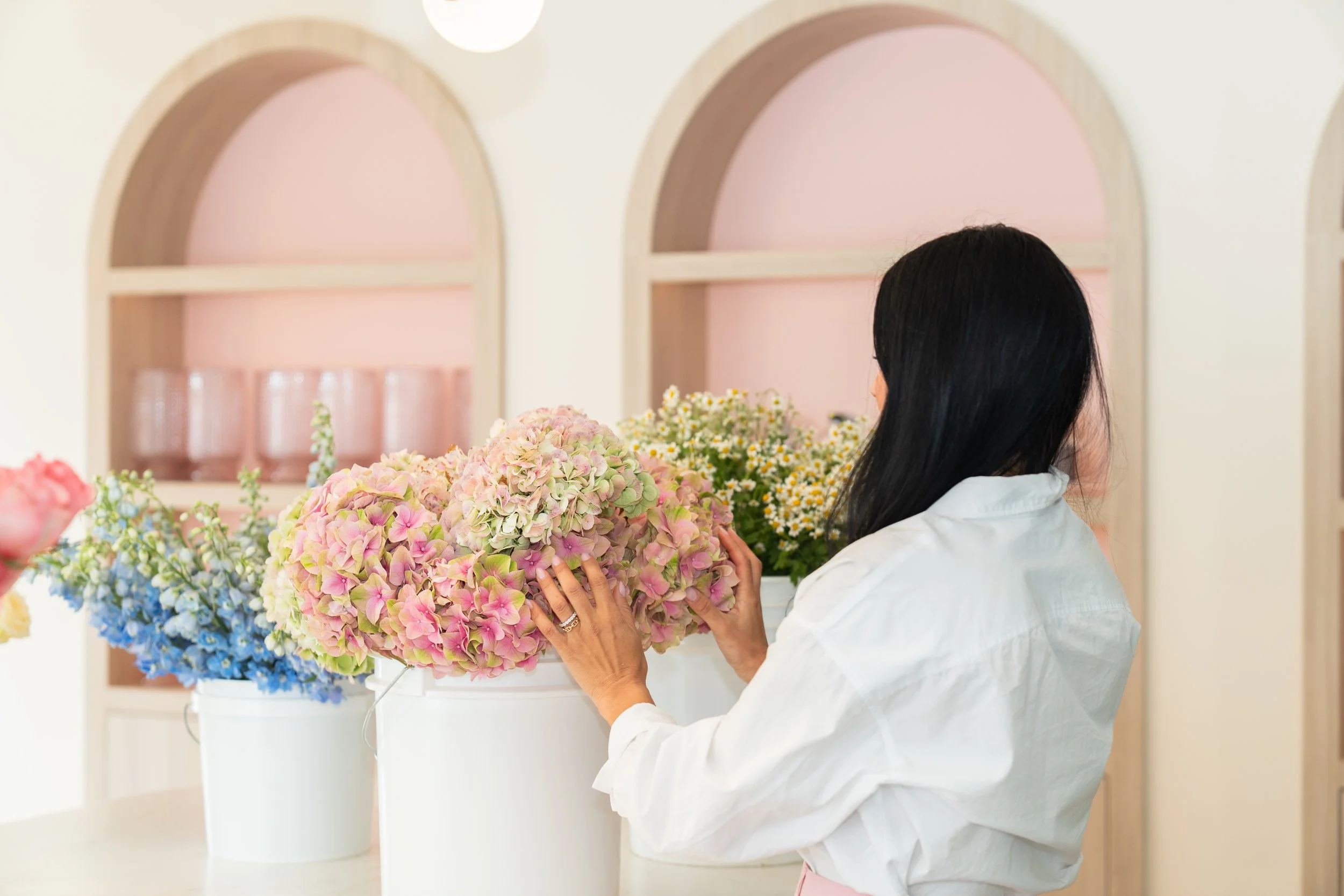 A woman arranging pink, white, and purple hydrangea flowers in front of a light pink and beige background.