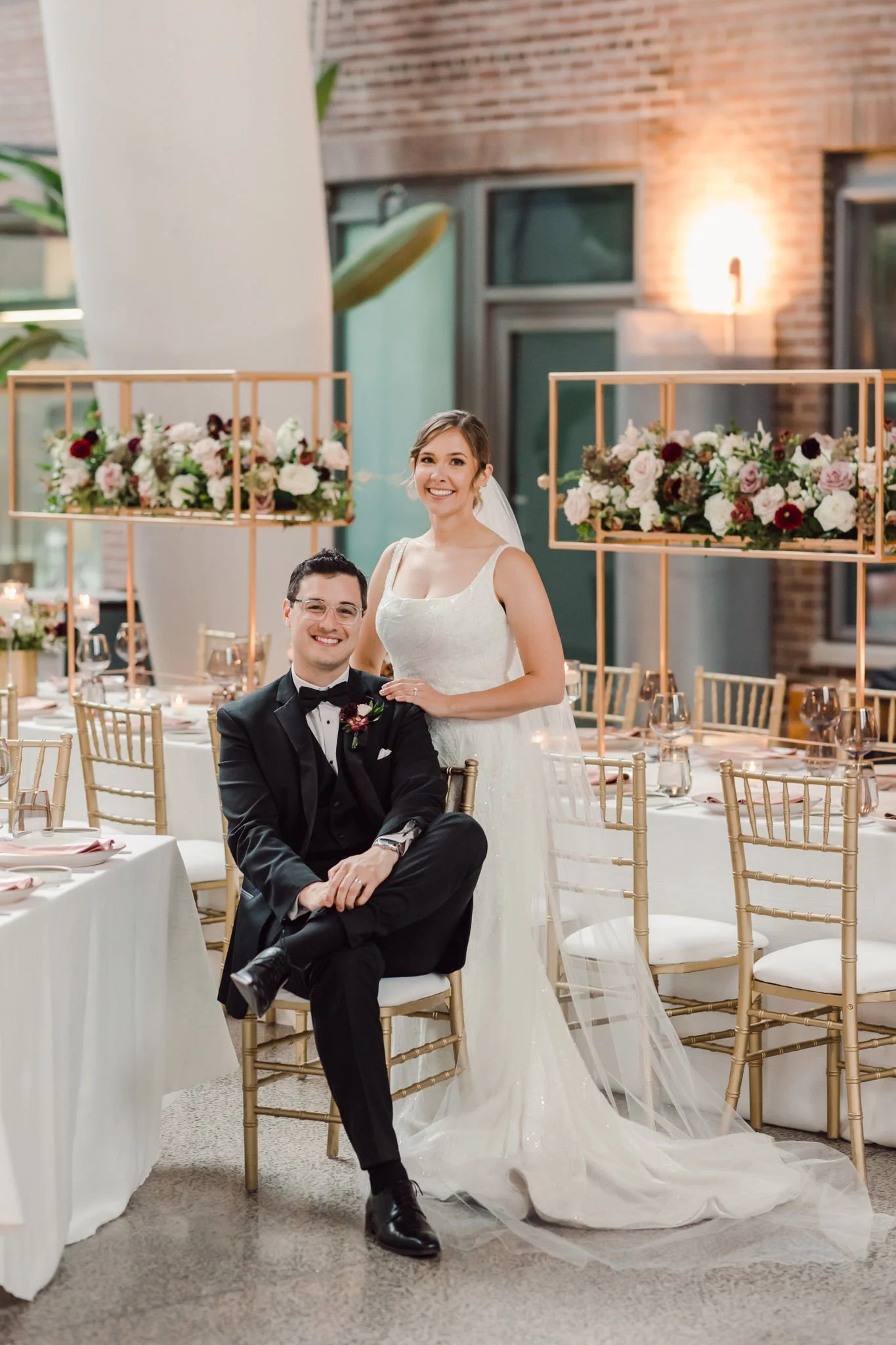 A bride and groom at a wedding reception, with the bride standing and smiling and the groom sitting on a chair, in a decorated banquet hall with floral centerpieces.