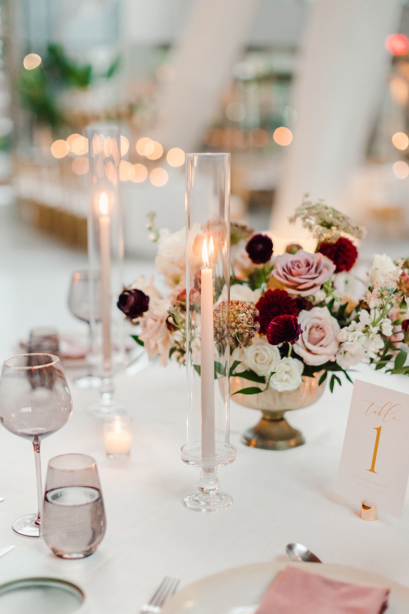Elegant wedding table setting with a floral centerpiece, tall candles in glass holders, wine glasses, and a table number card, set in a modern indoor venue.