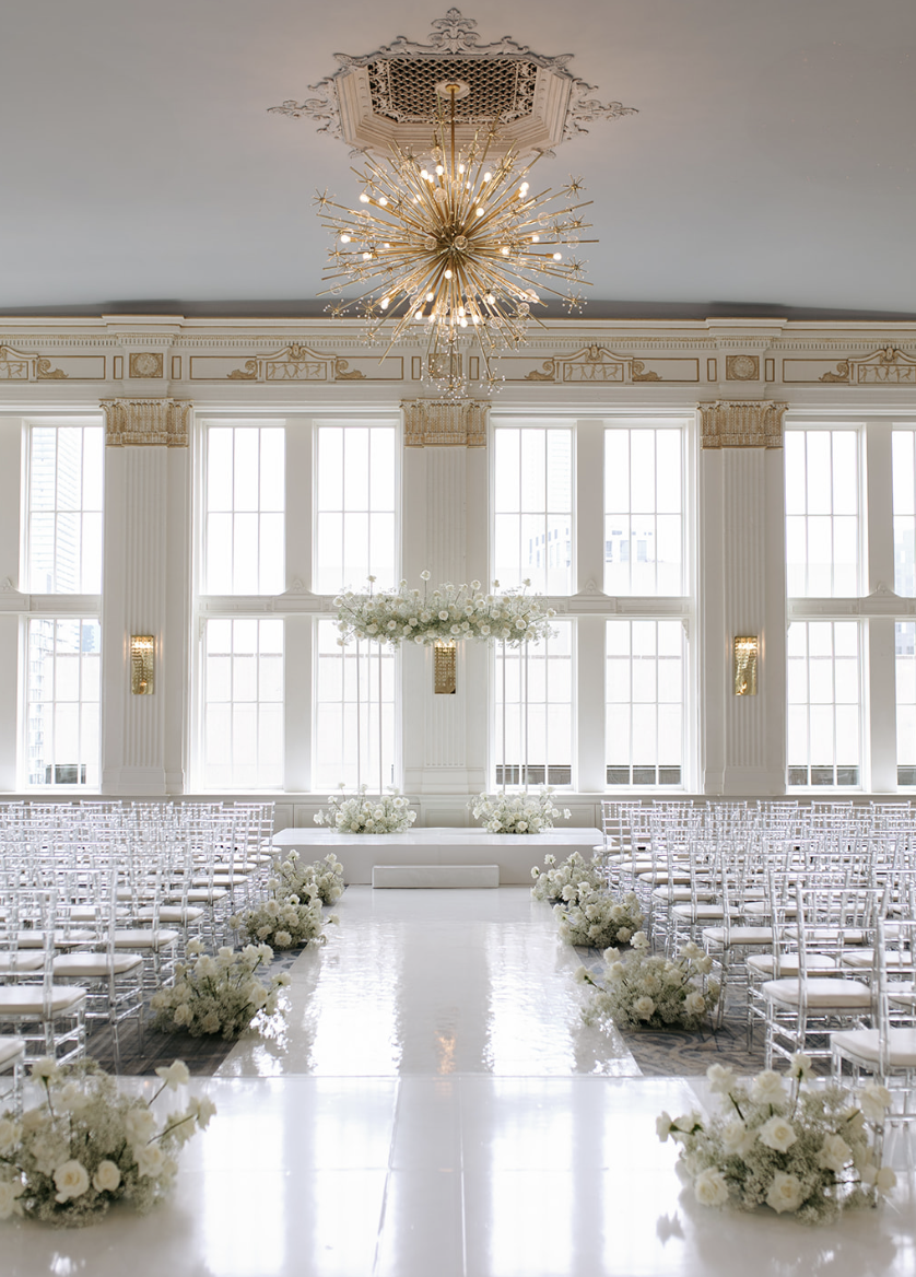 Elegant wedding ceremony setup in a bright room with tall windows, white floral arrangements, and rows of transparent chairs.
