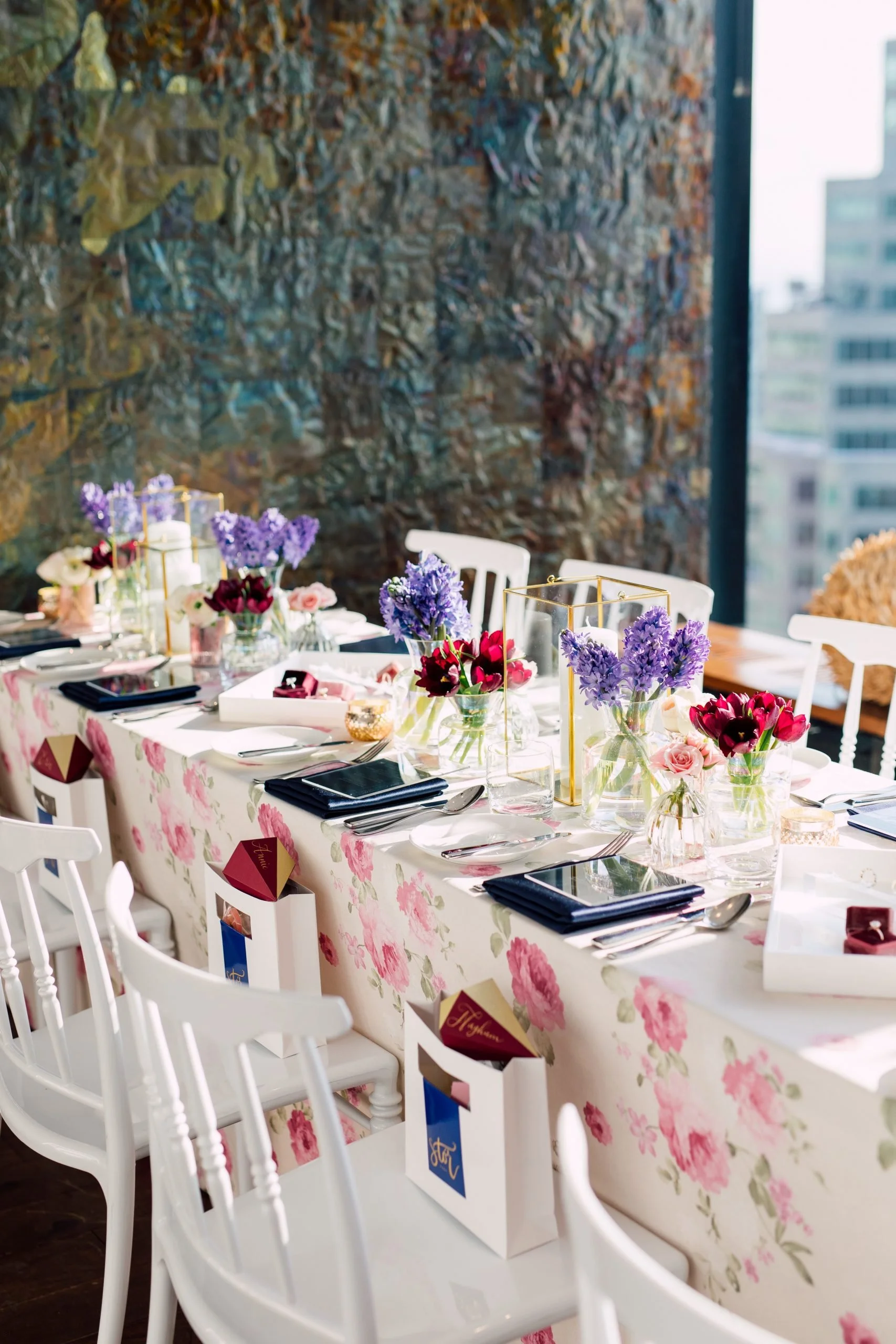 A beautifully decorated indoor dining table with a floral tablecloth, filled with vases of purple and red flowers, set with plates, utensils, and black napkins. There are small gift boxes on each chair and tall gold geometric candle holders, with city buildings visible through the windows in the background.