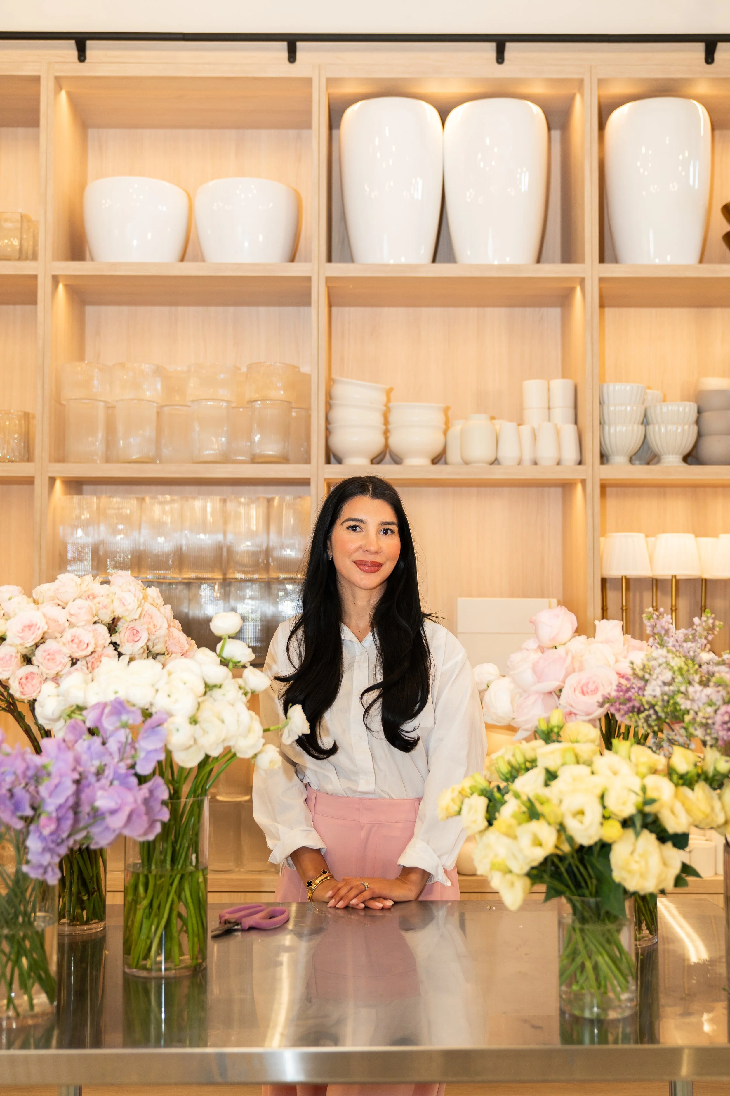 A woman standing behind a stainless steel table with colorful flowers in vases, in a well-lit store or flower shop with white shelves filled with white vases, bowls, and glassware in the background.