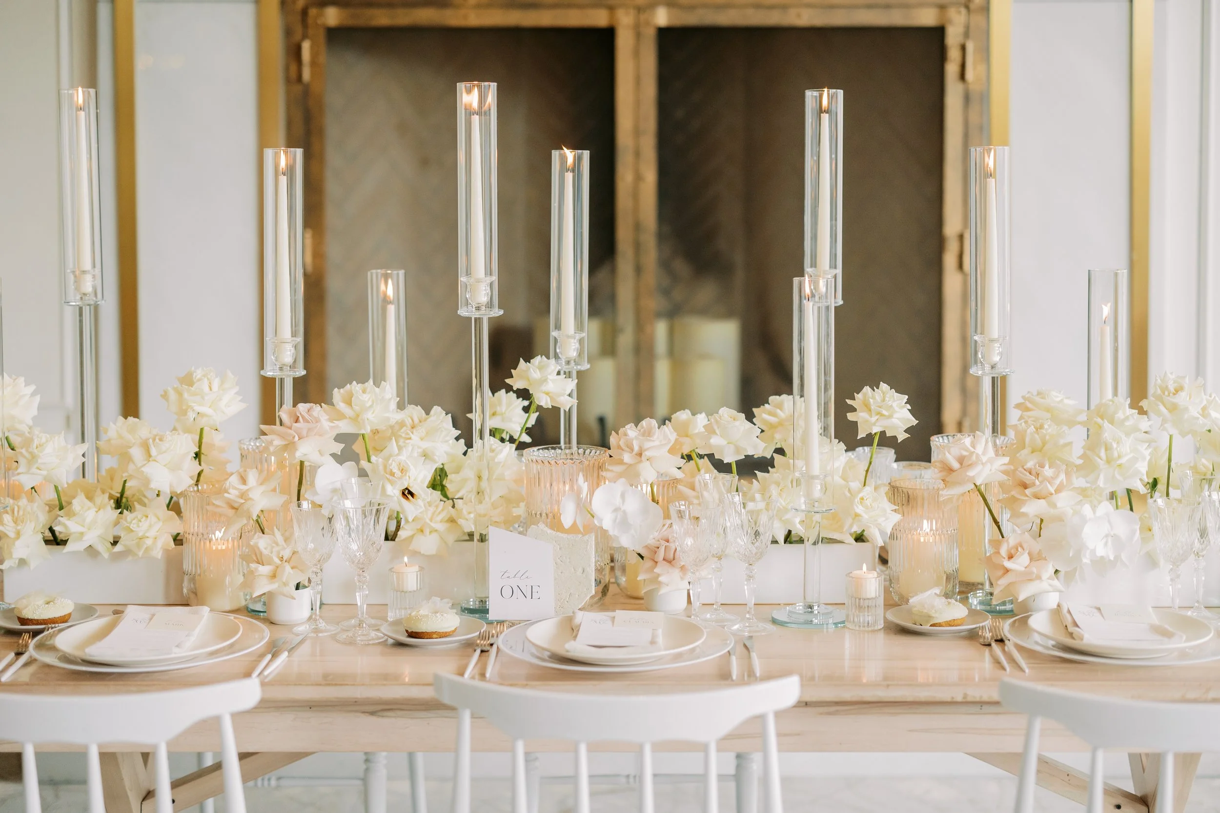 Elegant table centerpiece with white and blush flowers, tall glass candles, and table settings for a wedding or special event.