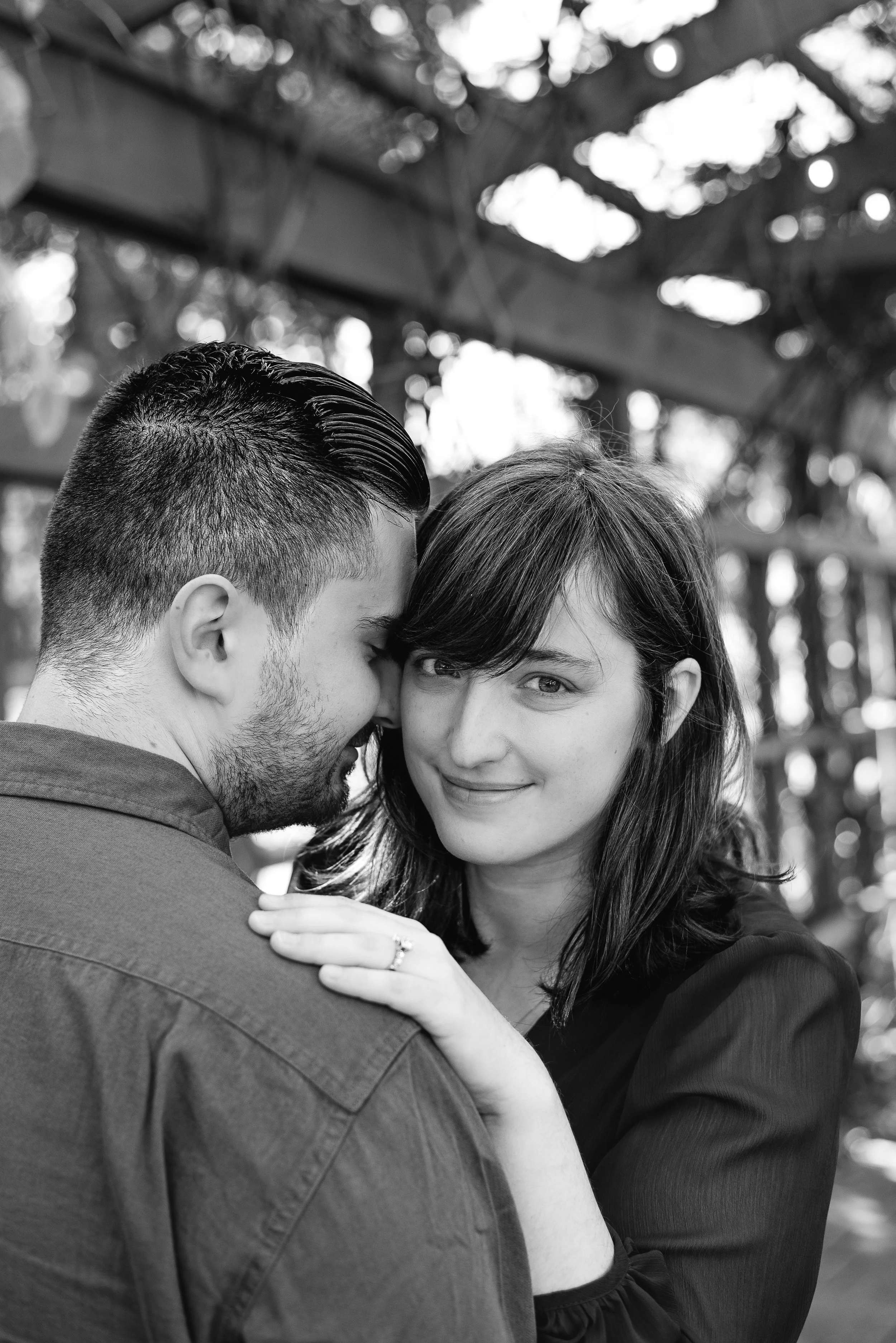 A black and white photo of a couple in an outdoor setting, with the man whispering into the woman's ear while she smiles and looks at the camera. She has her hand on his shoulder, displaying an engagement ring.