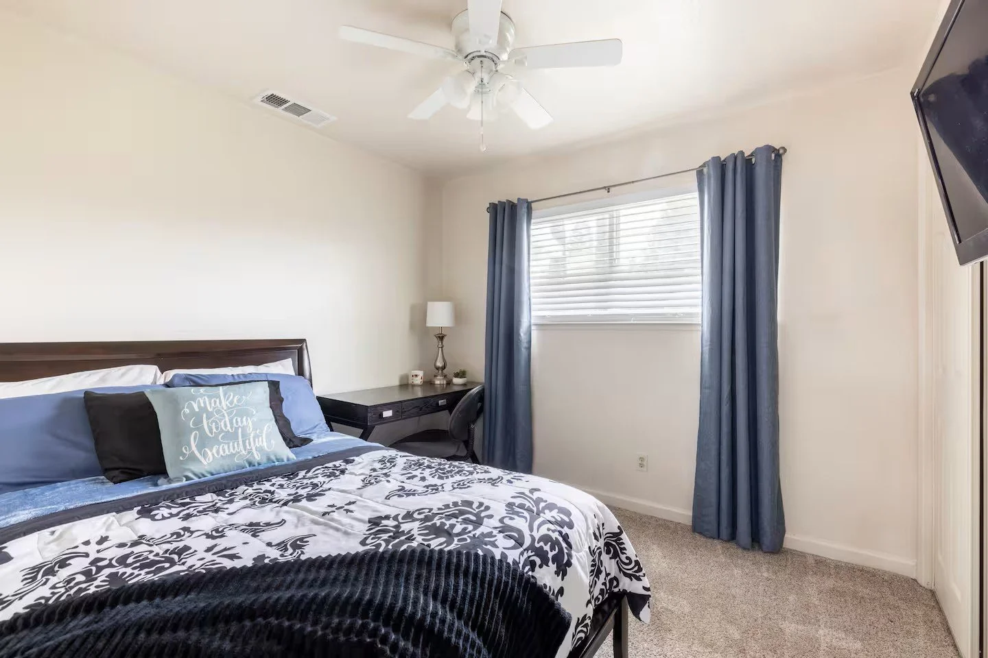Bedroom with a bed, pillows, window with blue curtains, ceiling fan, wall-mounted TV, and a desk with a lamp and chair.
