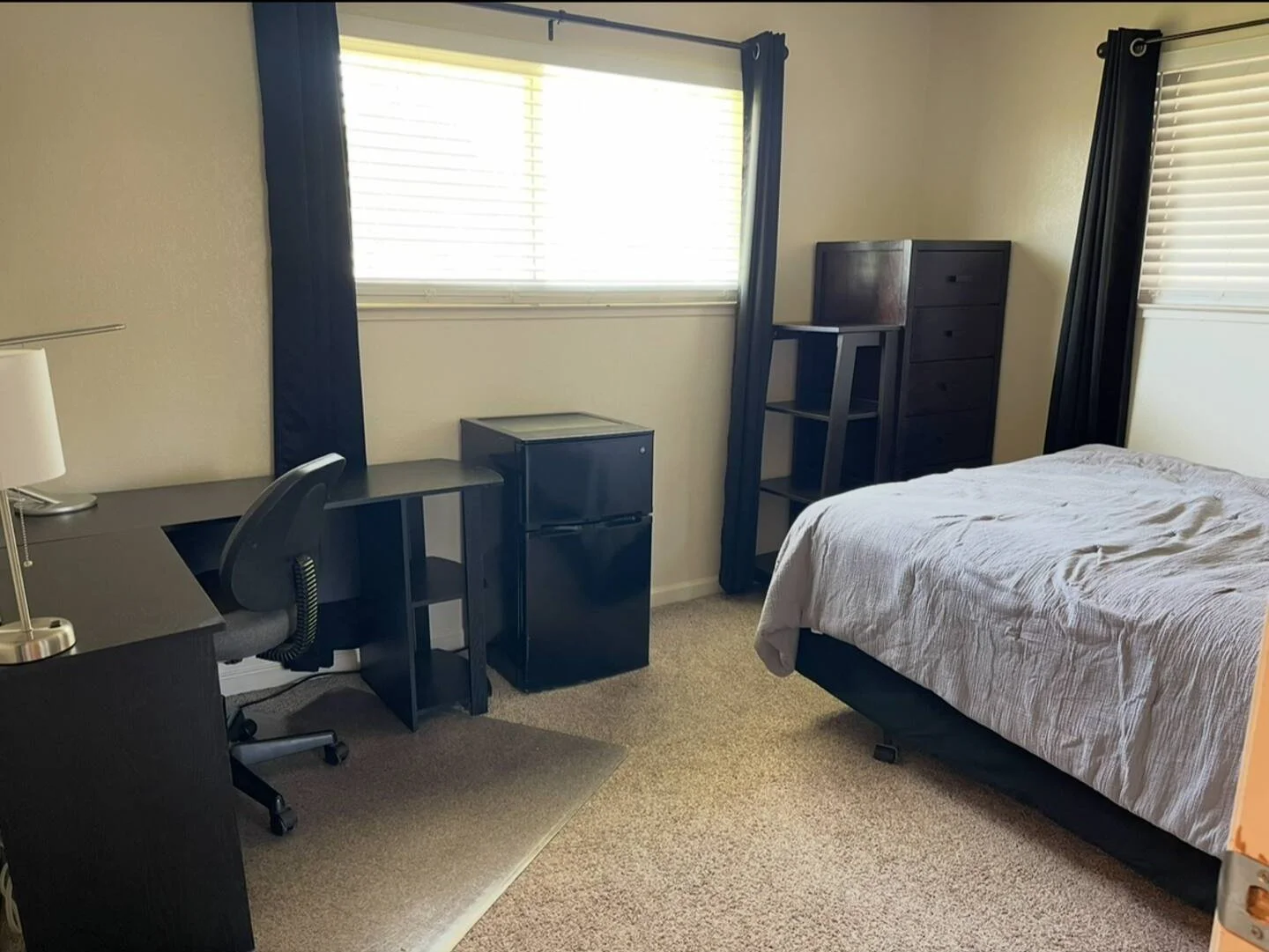 A bedroom with a bed, a desk with a lamp and office chair, a mini refrigerator, a chest of drawers, and a window with blinds and dark curtains.
