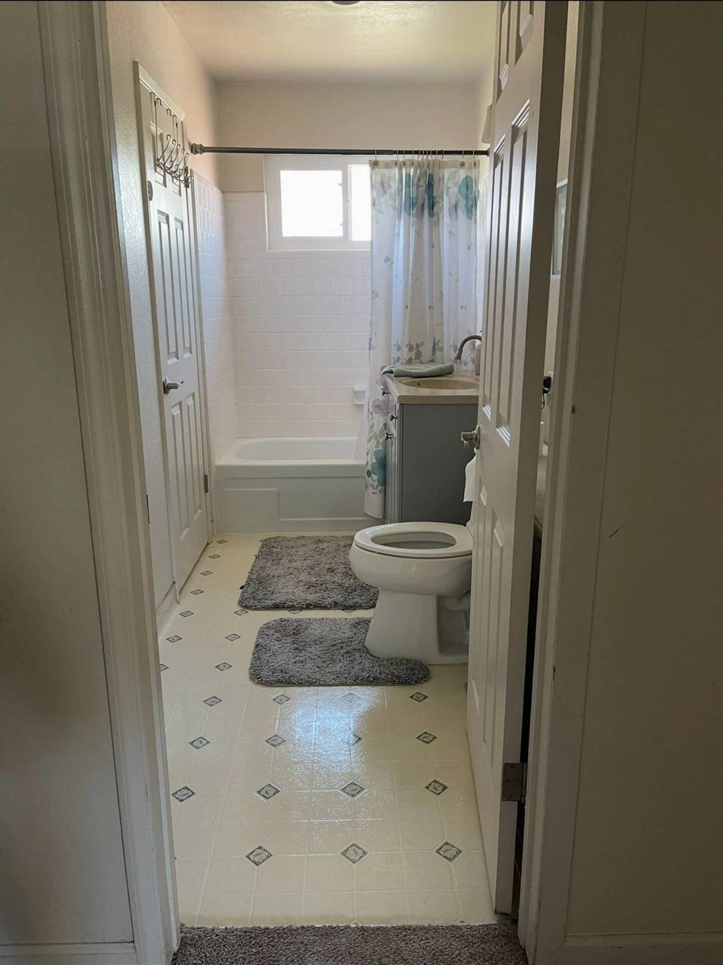 Interior of a bathroom showing a toilet with a space behind a door, a bathtub with a floral shower curtain, a small window, a sink with a cabinet, gray bathroom rugs, and beige tiled flooring.