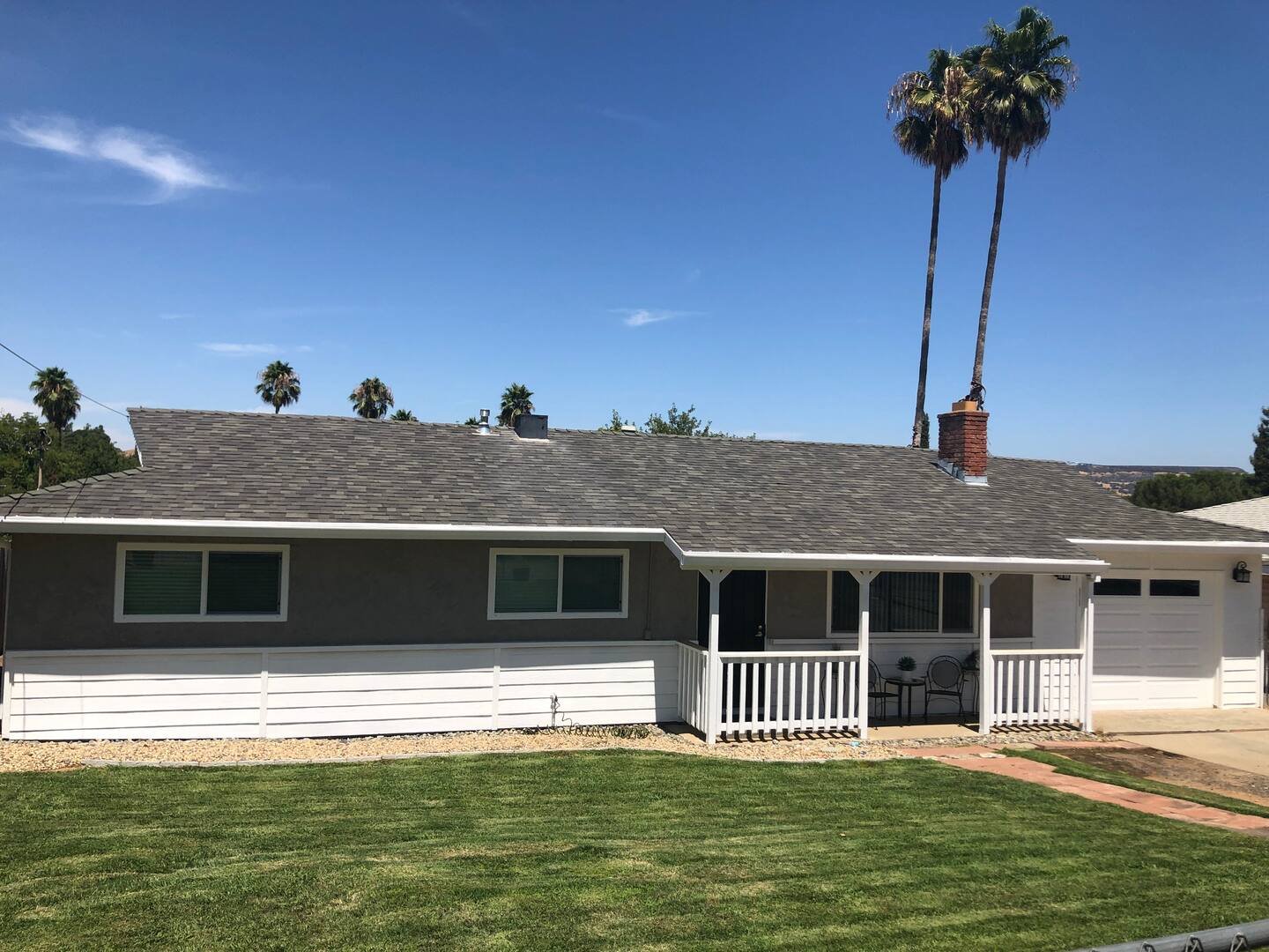 Front view of a single-story house with gray exterior walls, white trim, a small front porch with chairs, and a white garage door, set against a bright blue sky with palm trees in the background.