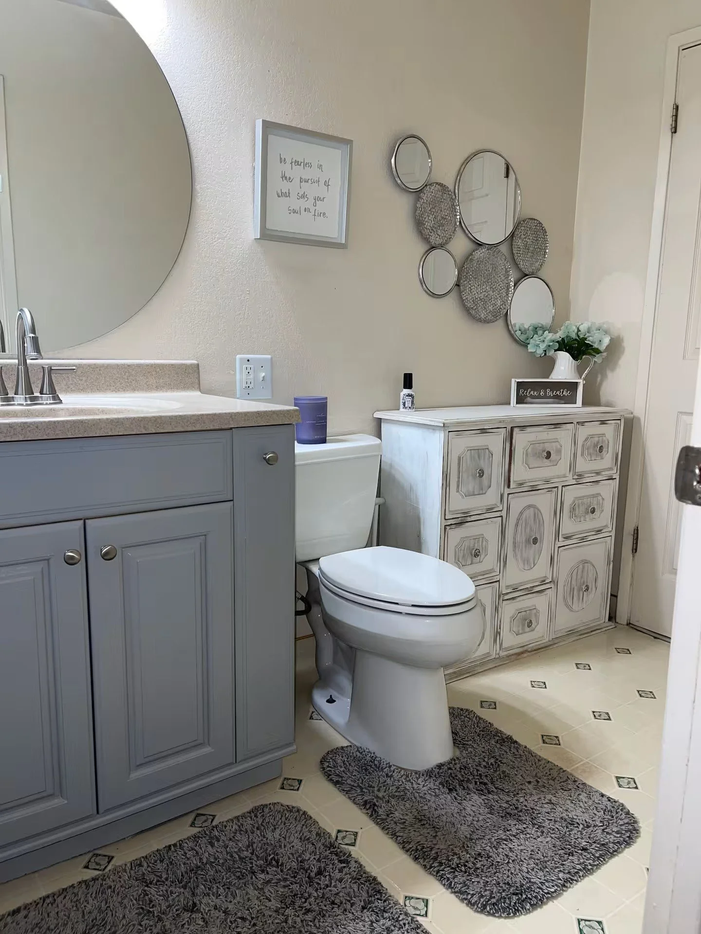 A bathroom with a gray vanity, a white toilet, a textured gray rug, a decorative cabinet, wall mirrors, and wall art.