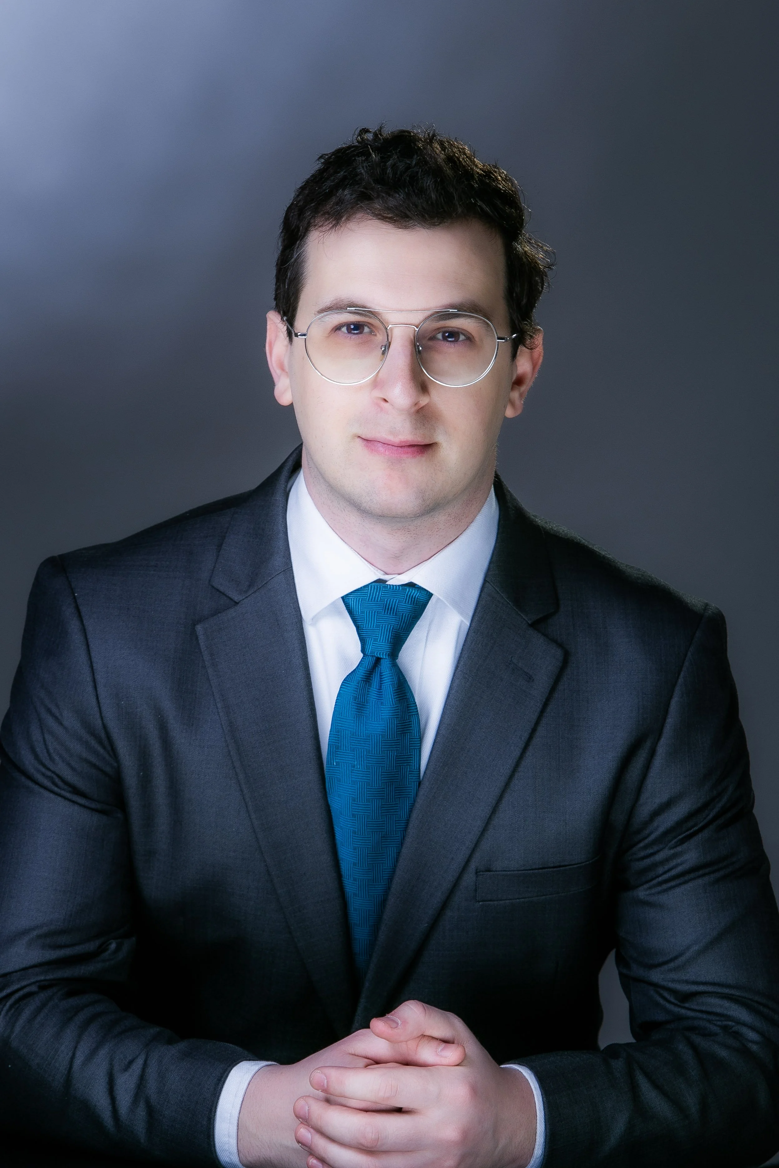 A man with glasses and dark hair wearing a suit, white shirt, and blue tie, sitting with hands clasped in front of a dark, gradient background.
