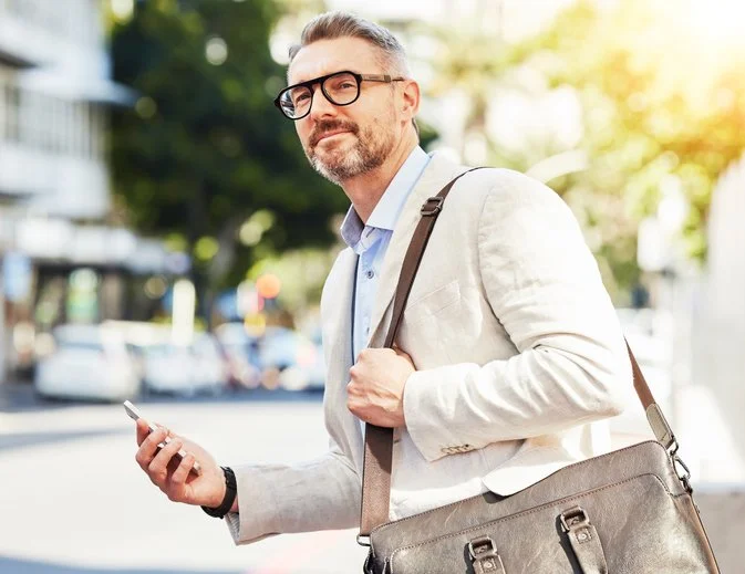 A middle-aged man with glasses and a beard standing outdoors on a city street, holding a smartphone in his right hand and carrying a brown shoulder bag, wearing a light-colored blazer over a blue shirt, with blurred cars and buildings in the background.
