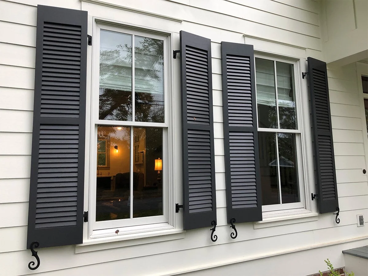 Two white-framed windows with black shutters on a white house exterior, with the view into the interior showing warm lighting and furniture.