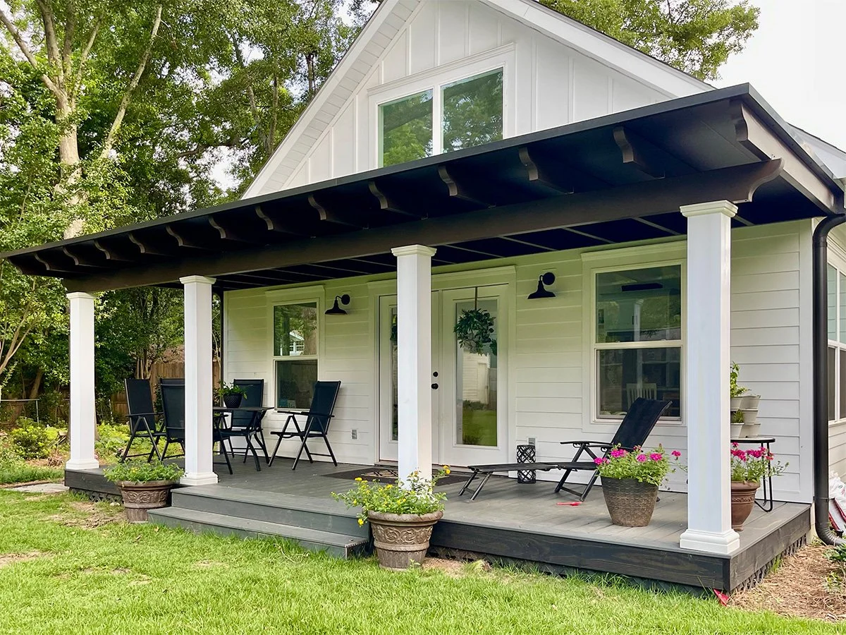 Back porch of a white house with black roof, black outdoor furniture, potted plants, and trees in the background.