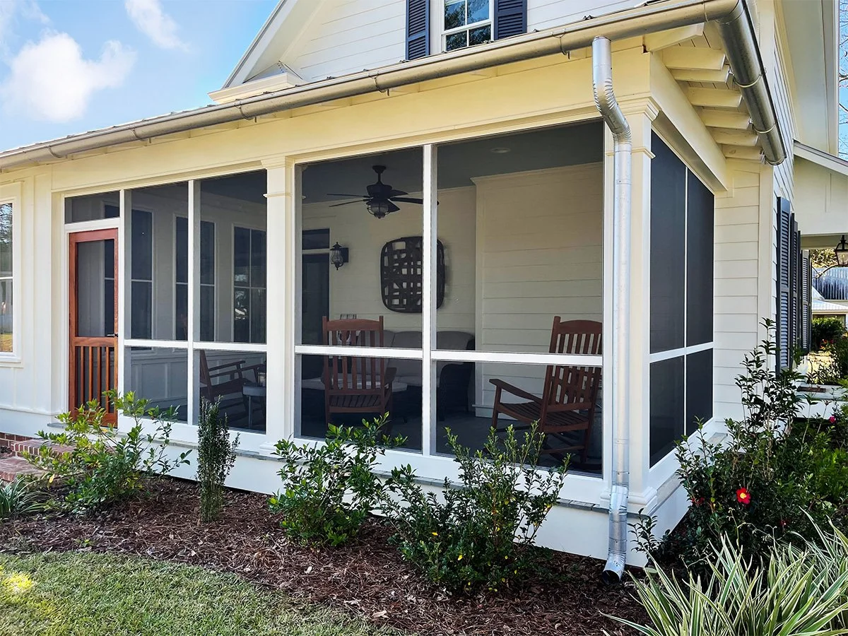 A screened-in porch attached to a house with four wooden chairs, a ceiling fan, and a decorative wall piece inside. The house has light-colored siding, black shutters, and a metal gutter system. There are shrubs and plants in front of the porch.