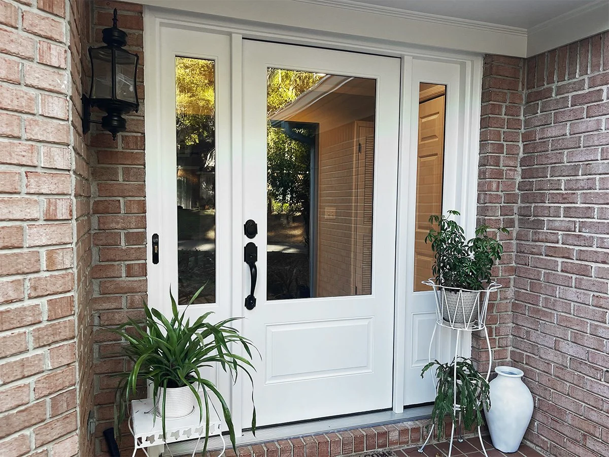 White front door with glass panels, brick wall, outdoor light fixture, potted plants, and a vase on a brick porch.