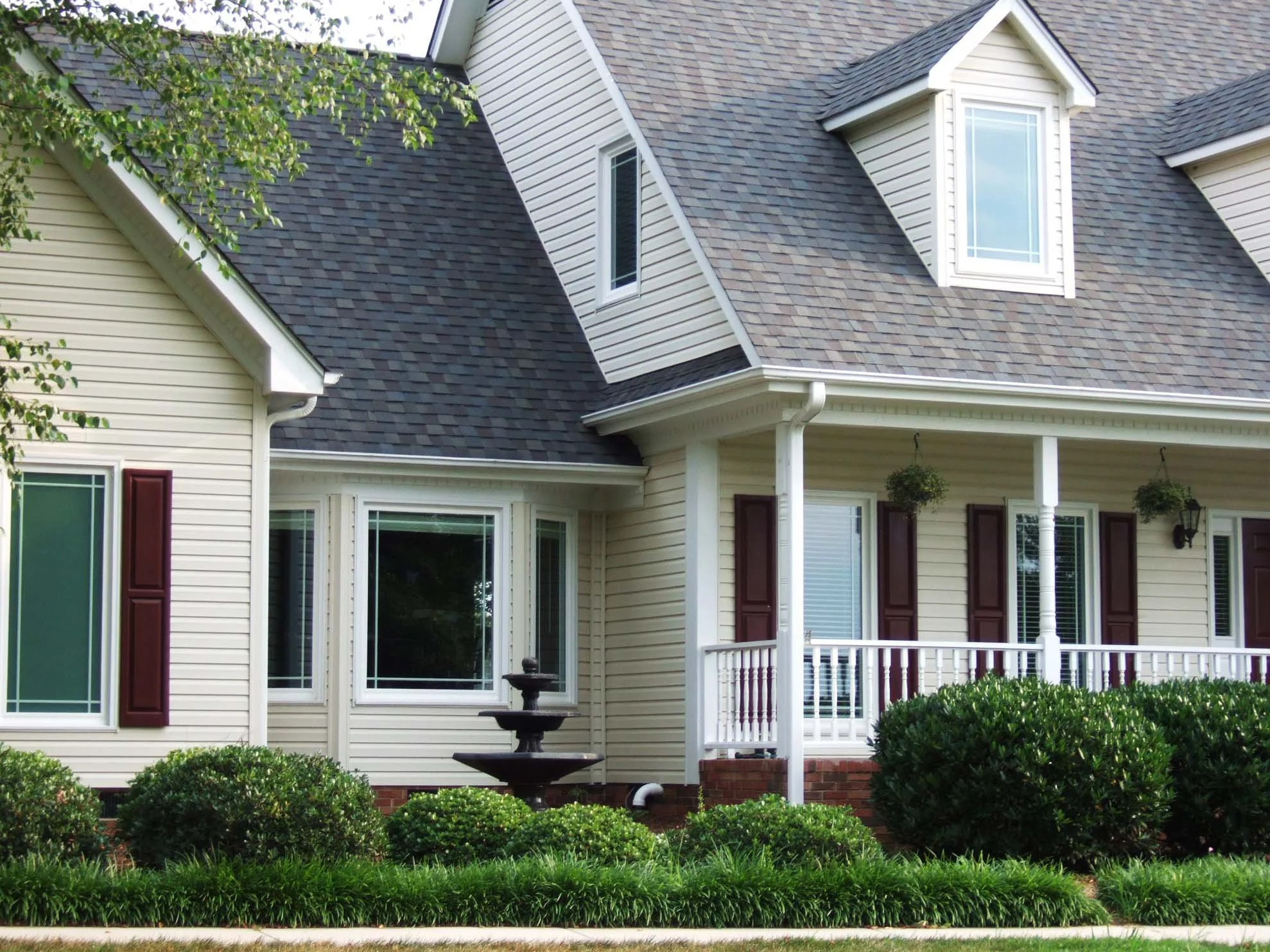 Front view of a house with beige vinyl siding, a multi-gabled roof with asphalt shingles, and a white porch. There are windows with white frames and burgundy shutters, a decorative birdbath in the front yard, and green bushes and trees surrounding the house.